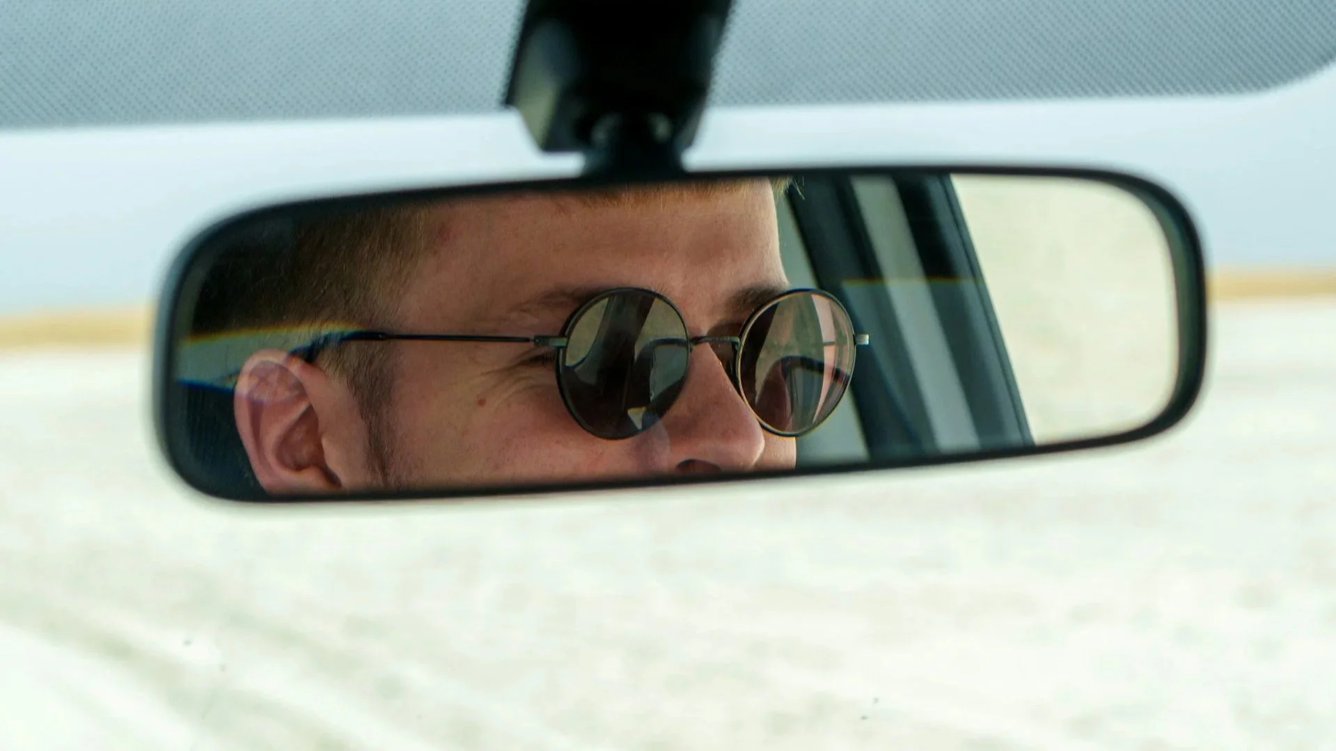 Close-up of a person wearing round sunglasses, looking at the road in the reflection of the rearview mirror.