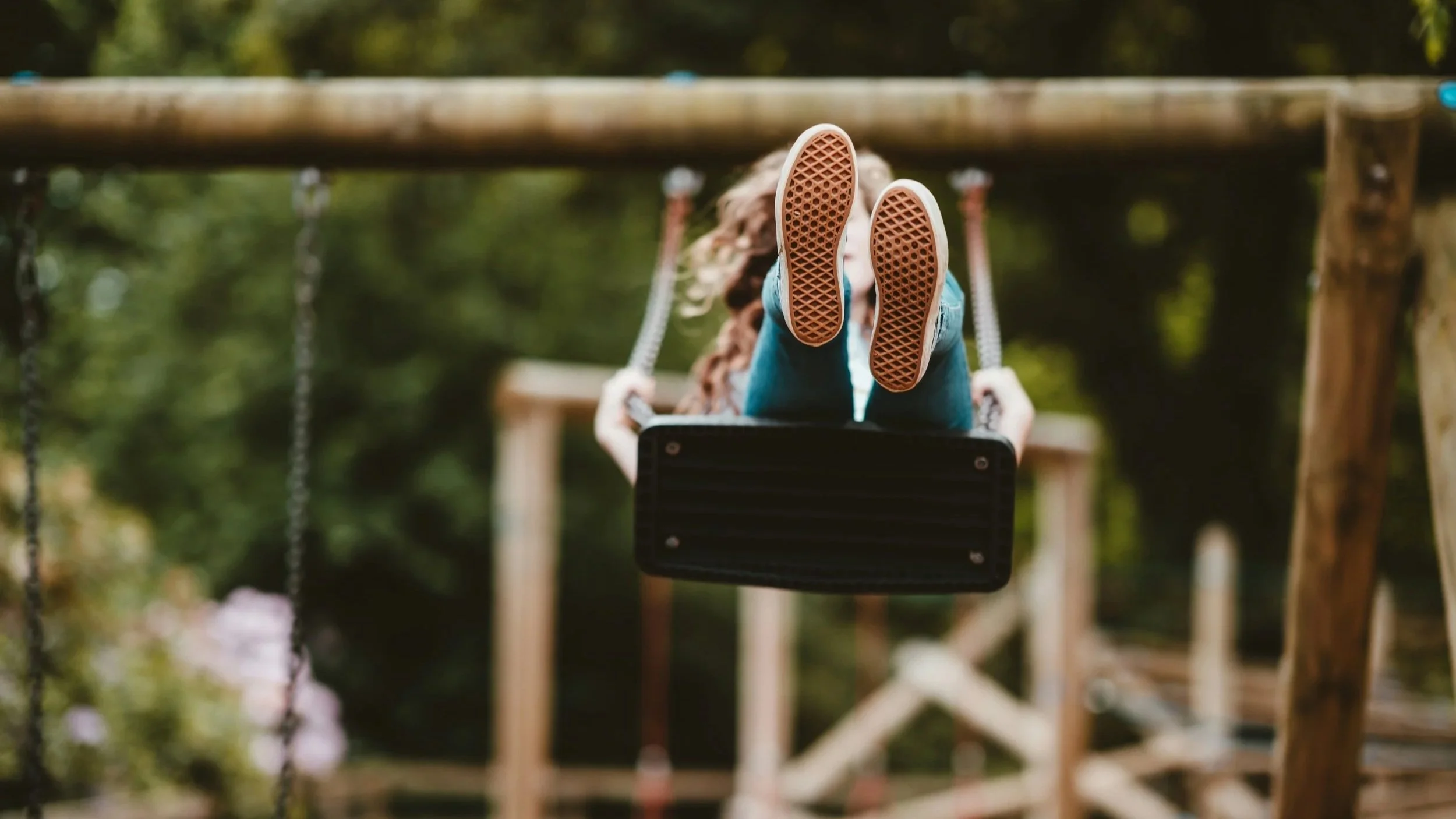A child on a swing in a park with trees in the background, facing away from the camera, wearing a light blue shirt and jeans.