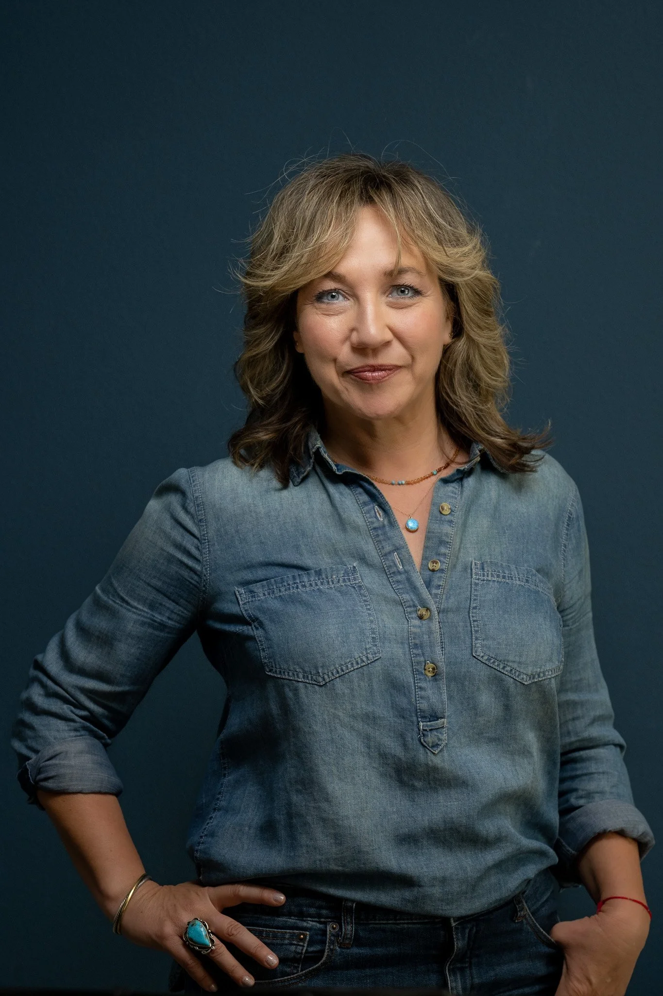 A woman with wavy, shoulder-length blonde hair, wearing a denim shirt with rolled-up sleeves, multiple necklaces, and jewelry on her fingers and wrist, standing against a dark blue background.