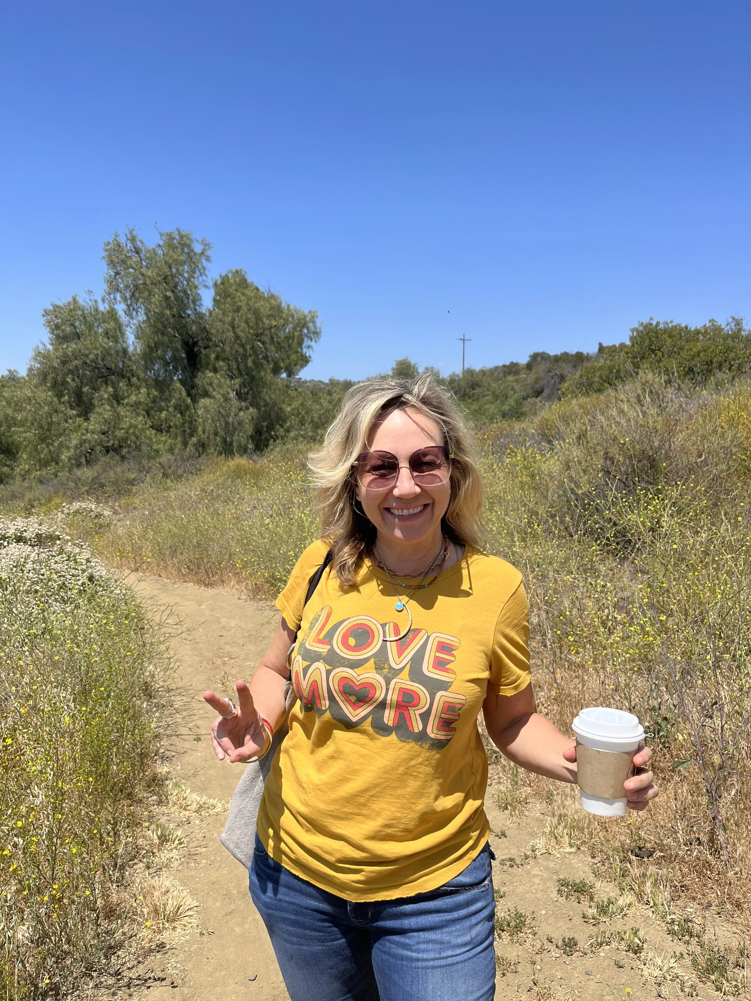 A woman smiling and making a peace sign with her right hand, holding a coffee cup in her left hand, standing on a trail in a field with bushes and trees under a clear blue sky.