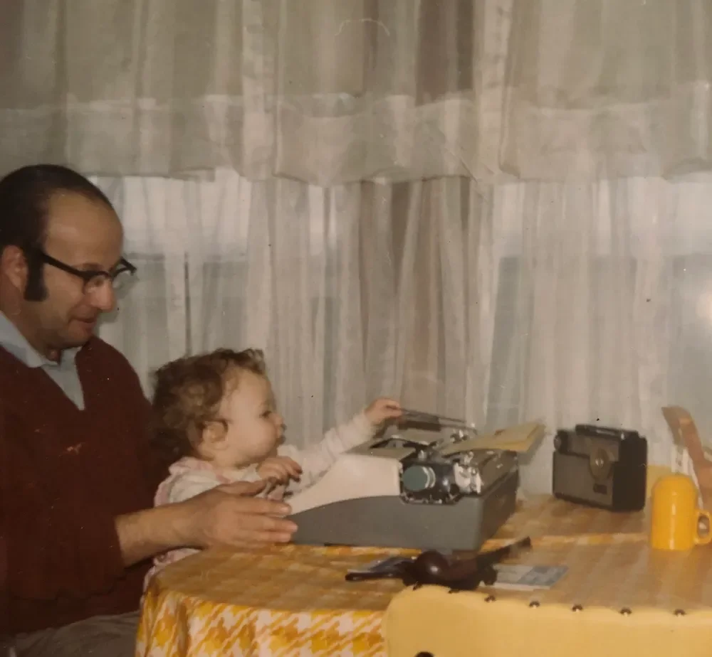 Man helping a young girl operate a vintage typewriter at a table with a yellow tablecloth, with old radio and a yellow mug in the background.