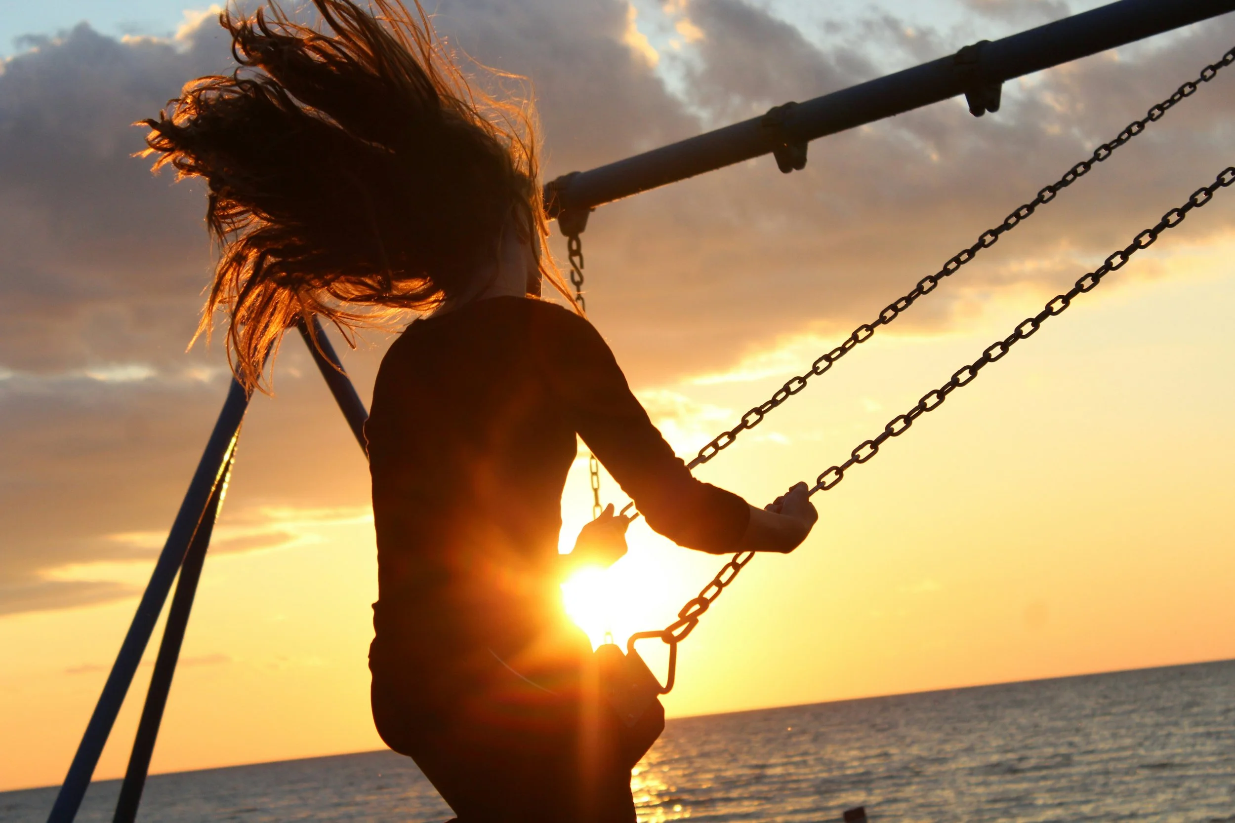 A child with flowing hair playing on a swing at sunset by the water.