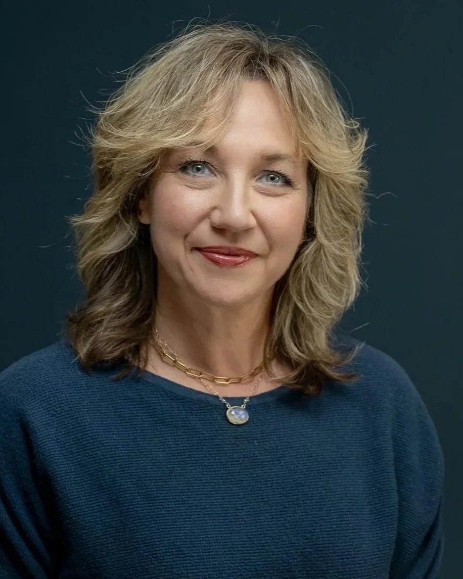 A portrait of a middle-aged woman with wavy blonde hair, blue eyes, and a warm smile, wearing a blue sweater and a gold necklace with a pendant, against a dark background.