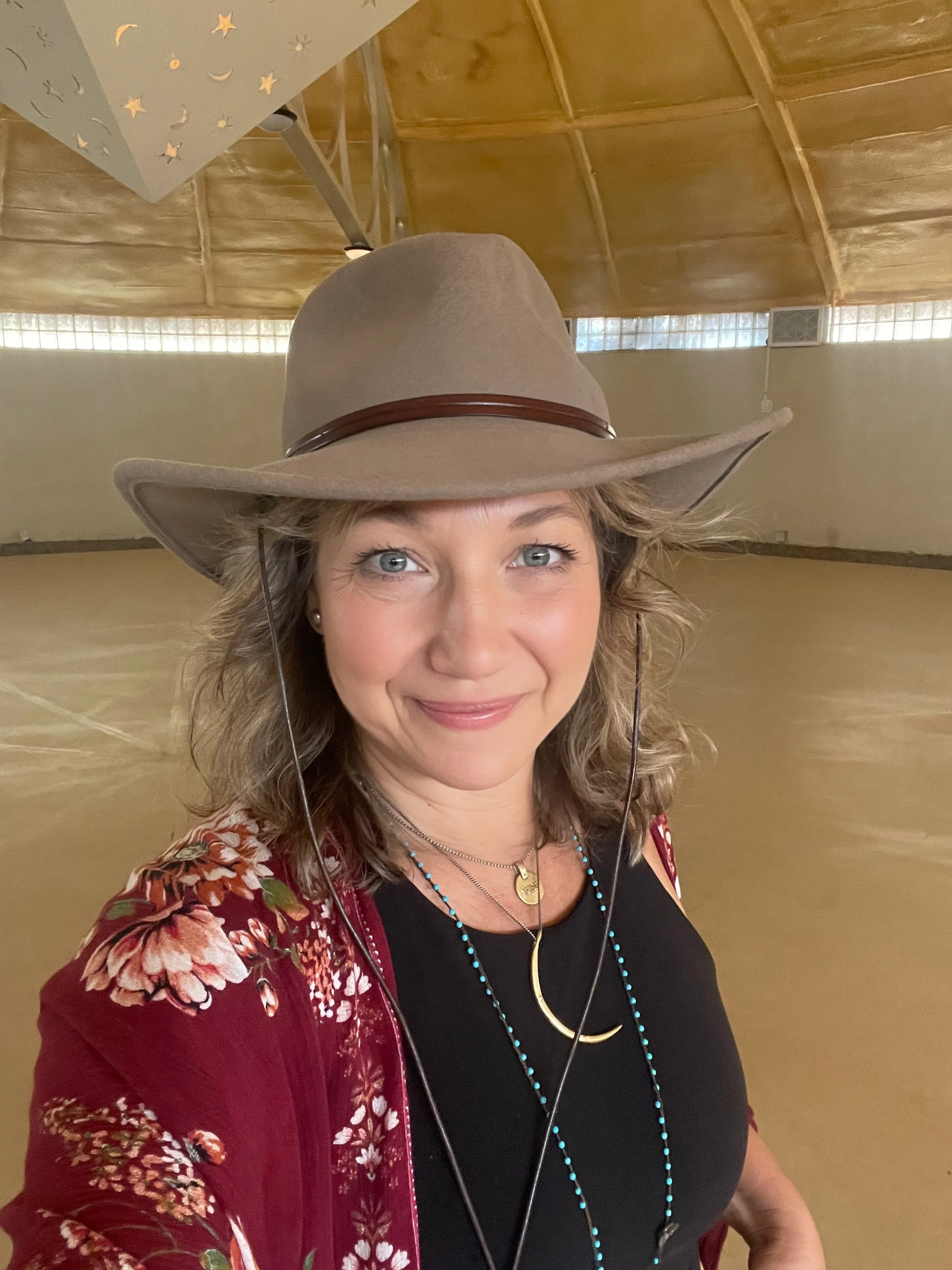 A woman with curly blonde hair wearing a wide-brimmed tan hat, black top, red floral jacket, and multiple necklaces, smiling indoors with a wooden ceiling and glass brick wall in the background.