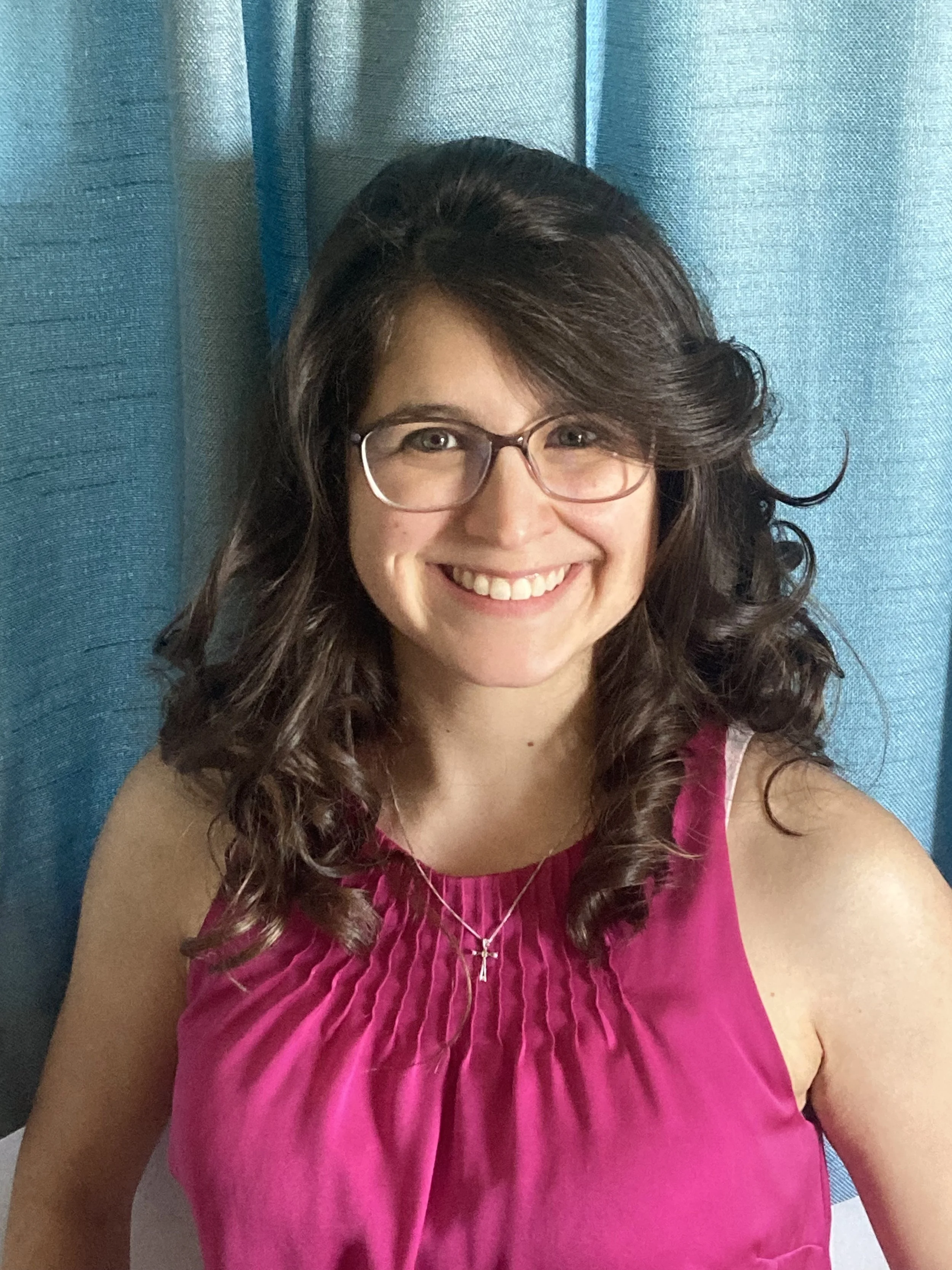 A smiling woman with dark curly hair wearing glasses and a pink ruffled sleeveless top, sitting in front of blue curtains.