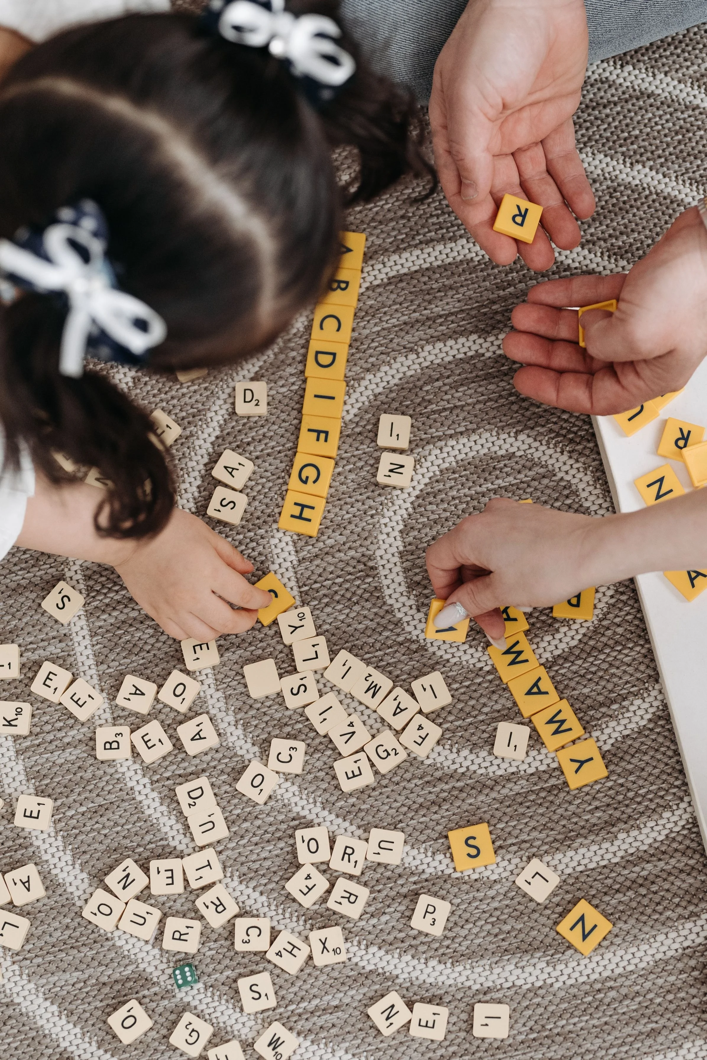Two children with pigtails and bows playing a word game with Scrabble tiles on a textured fabric surface, with scattered tiles and a die.