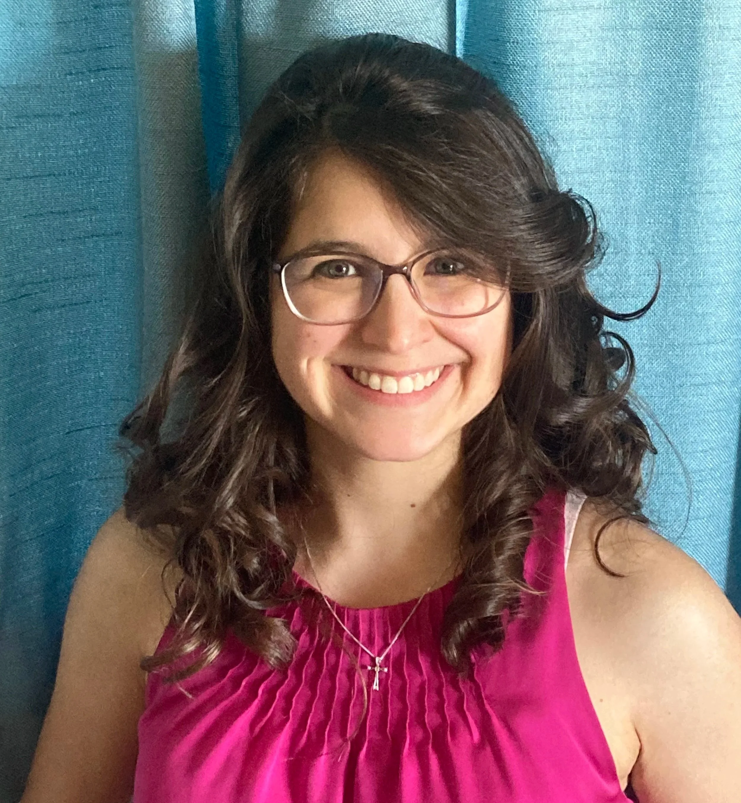 Close-up of a woman with curly brown hair and glasses, smiling, wearing a pink dress, against a blue background.