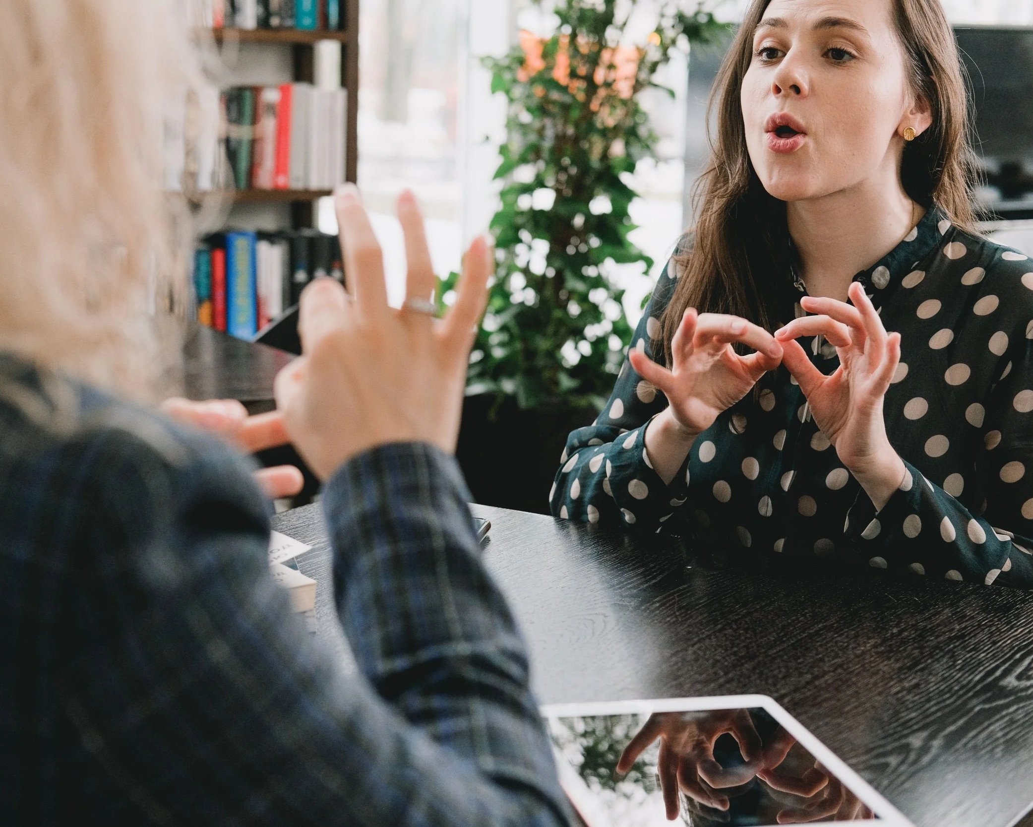 A woman with long brown hair, wearing a black dress with white polka dots, is speaking and gesturing with her hands during a conversation at a table in a bright room with large windows, green plants, and a bookshelf in the background.