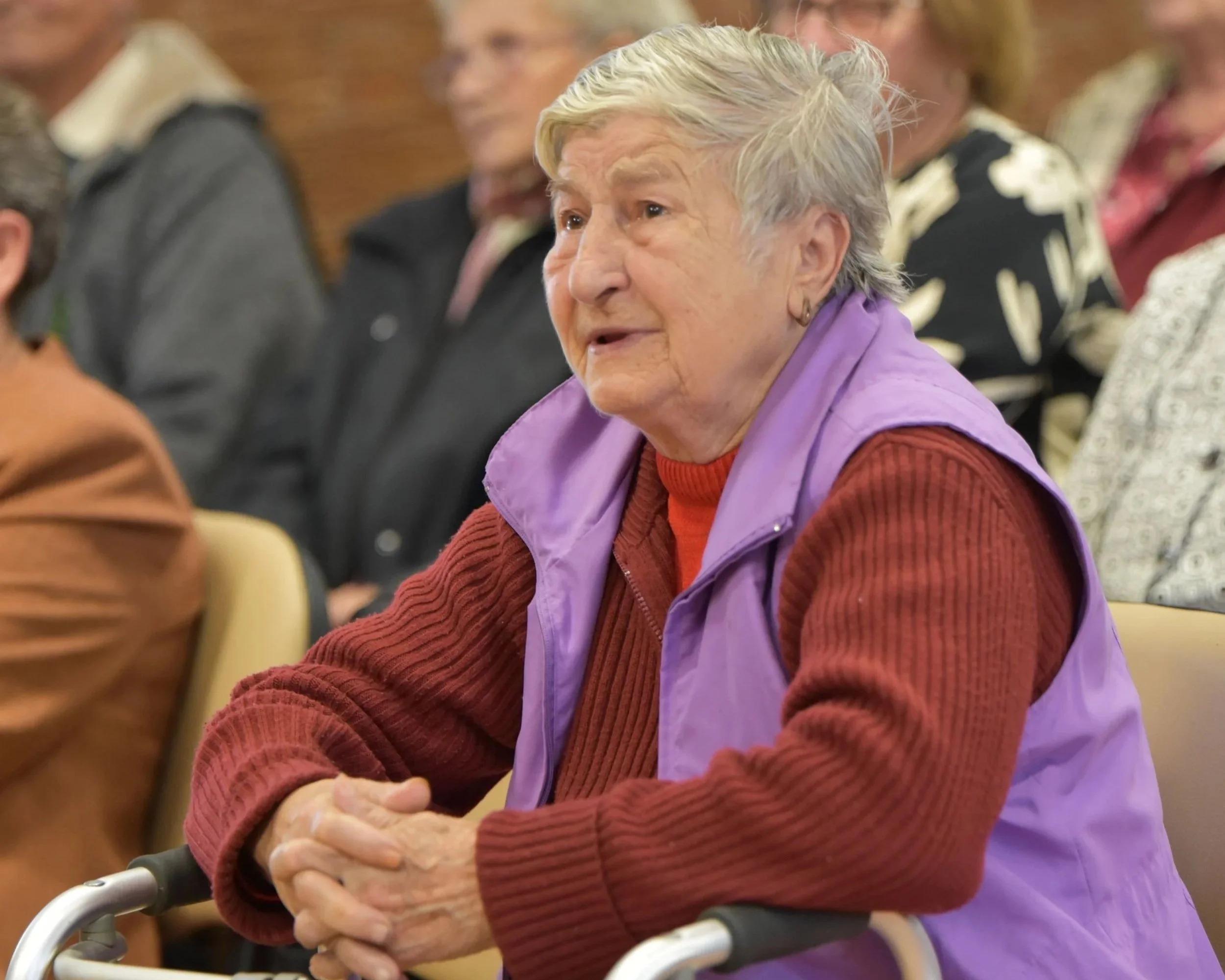 Older woman with gray hair wearing a purple vest and red sweater sitting at a table with her arms crossed, accompanied by other elderly individuals in the background in a social or community setting.