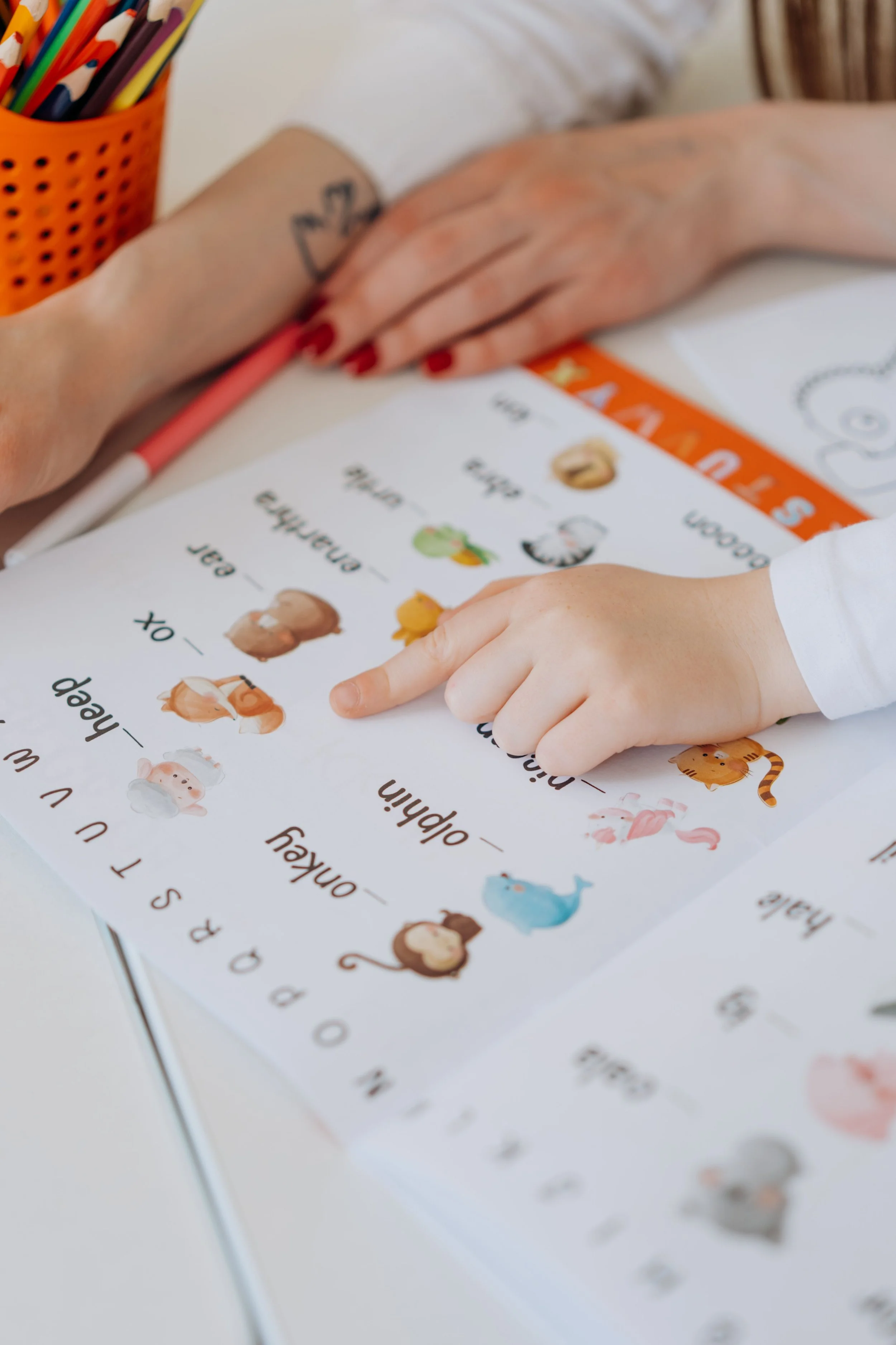 Close-up of a child's hand pointing to a worksheet with pictures of animals and words, with an adult's hand resting nearby. A container of colored pencils is visible in the background.