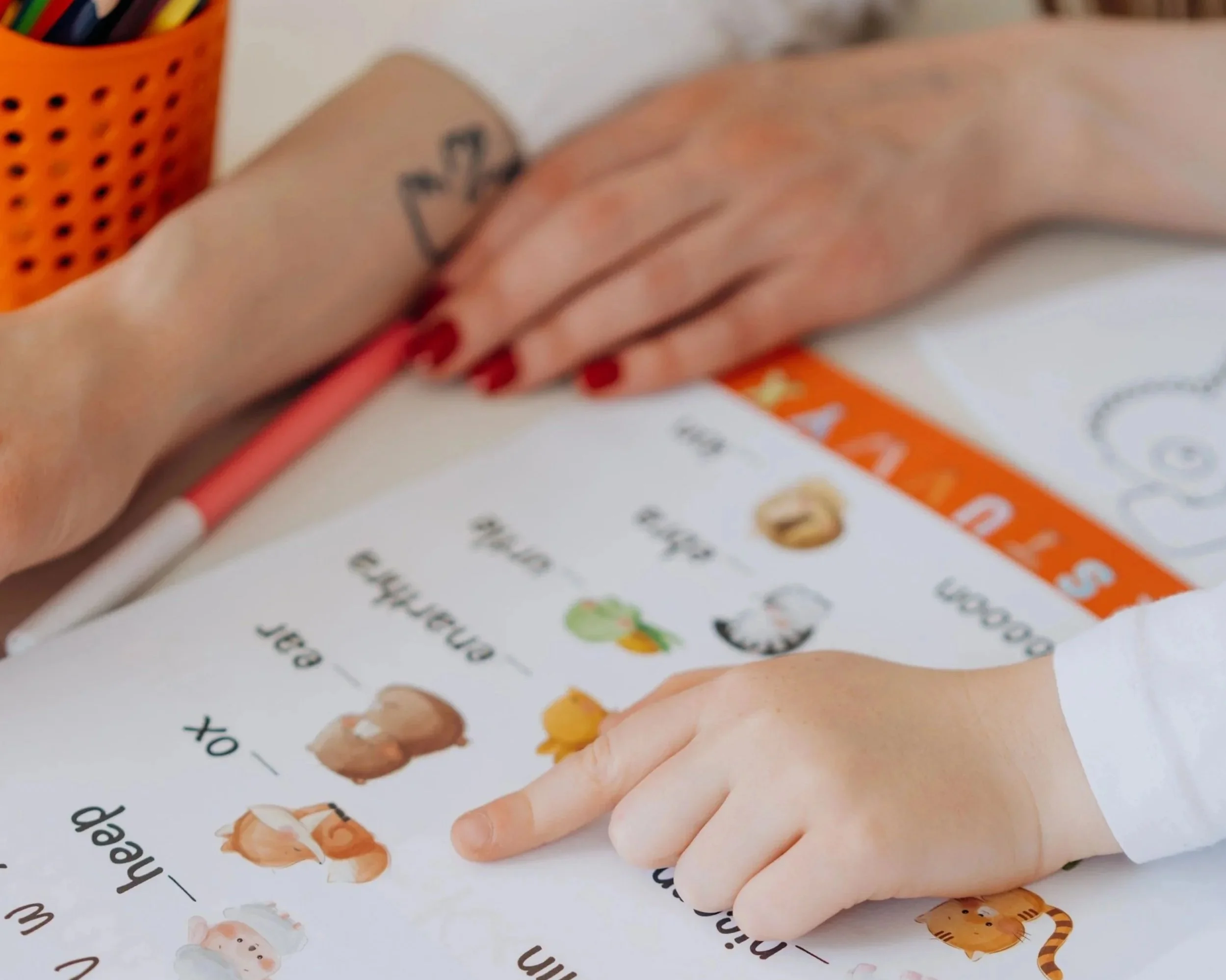 Close-up of a child's hand pointing to a worksheet with pictures of animals and words, with an adult's hand resting nearby. A container of colored pencils is visible in the background.