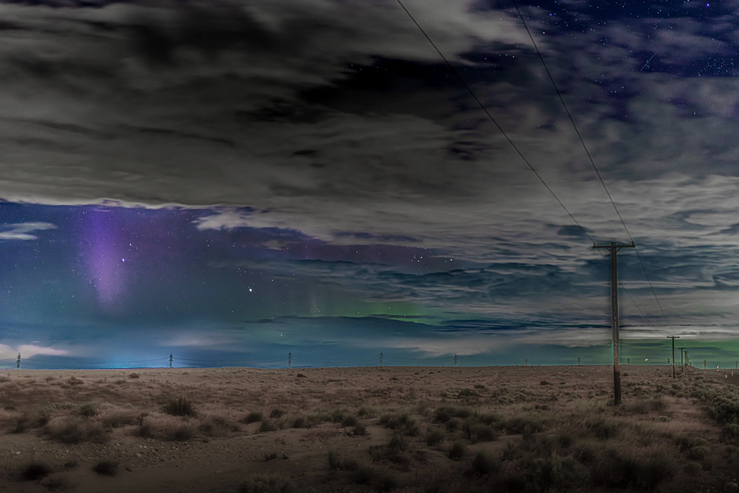 Thick clouds cross a starry night sky above open scrubland and a line of power poles north of Richland, Washington.