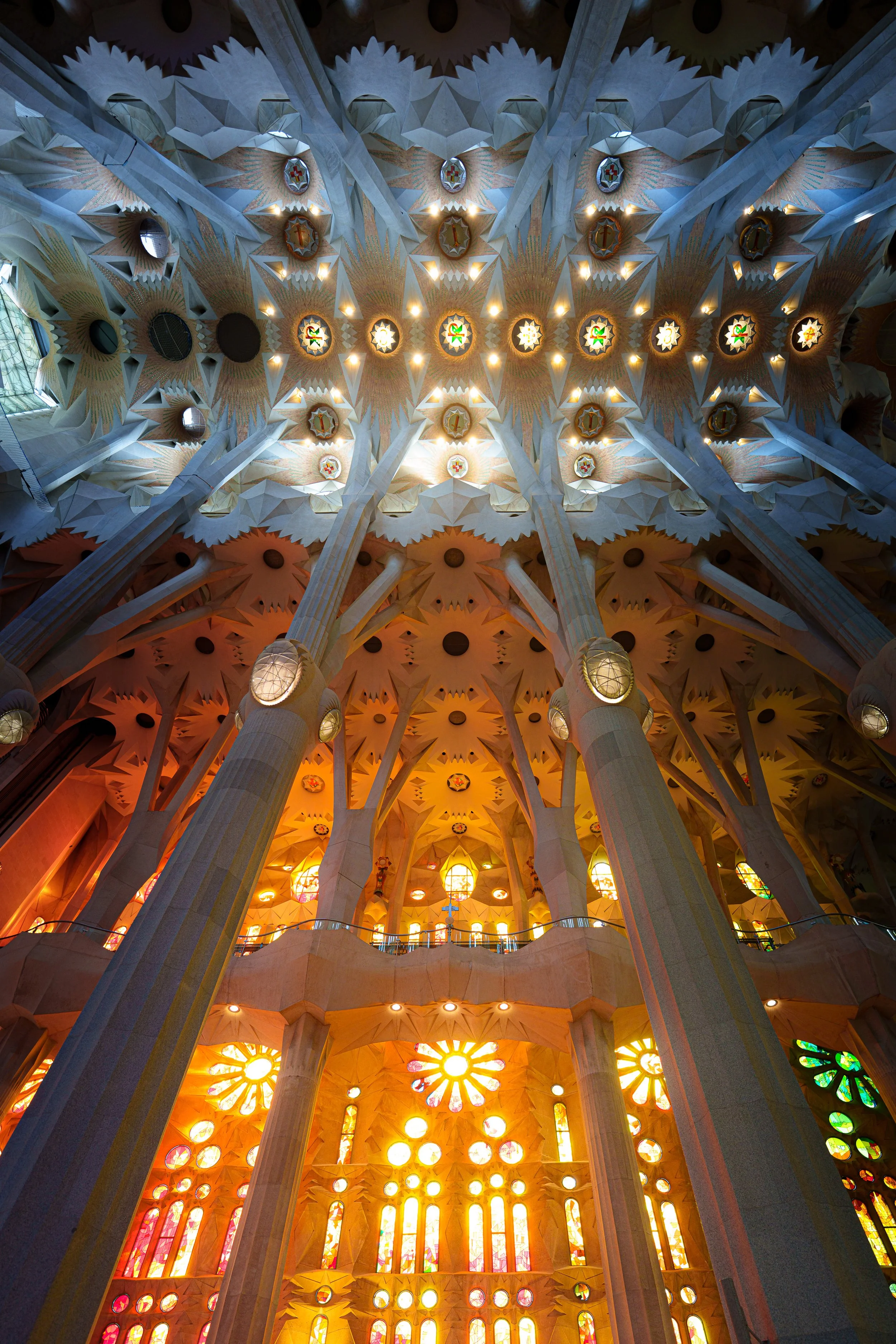Looking up at the ceiling of Sagrada Familia, showcasing detailed stained glass windows, tall stone columns, and intricate architectural designs in warm and cool lighting.