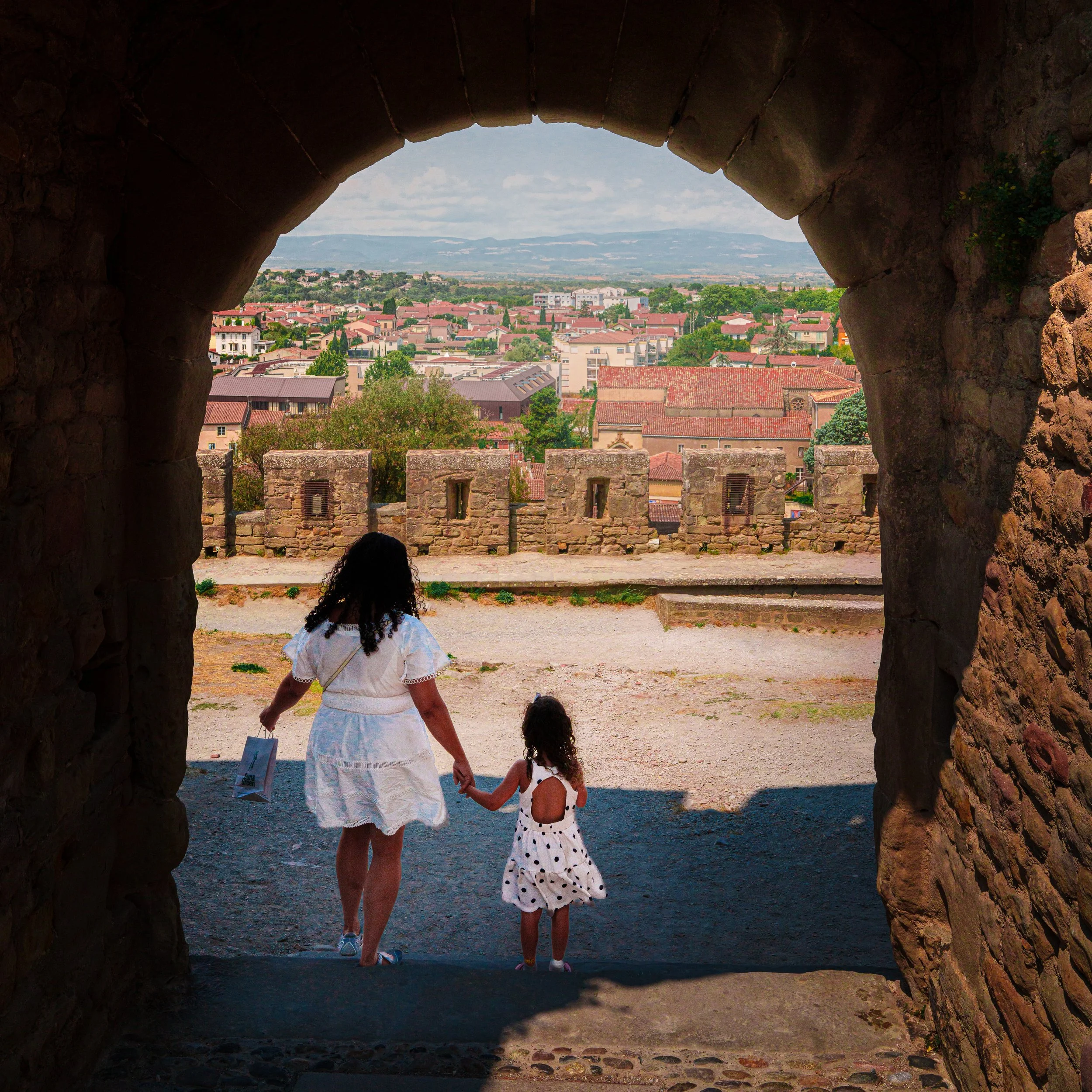 View of a woman and a girl holding hands as they walk through an ancient stone archway, overlooking a town with red-tiled roofs and distant mountains in the background.