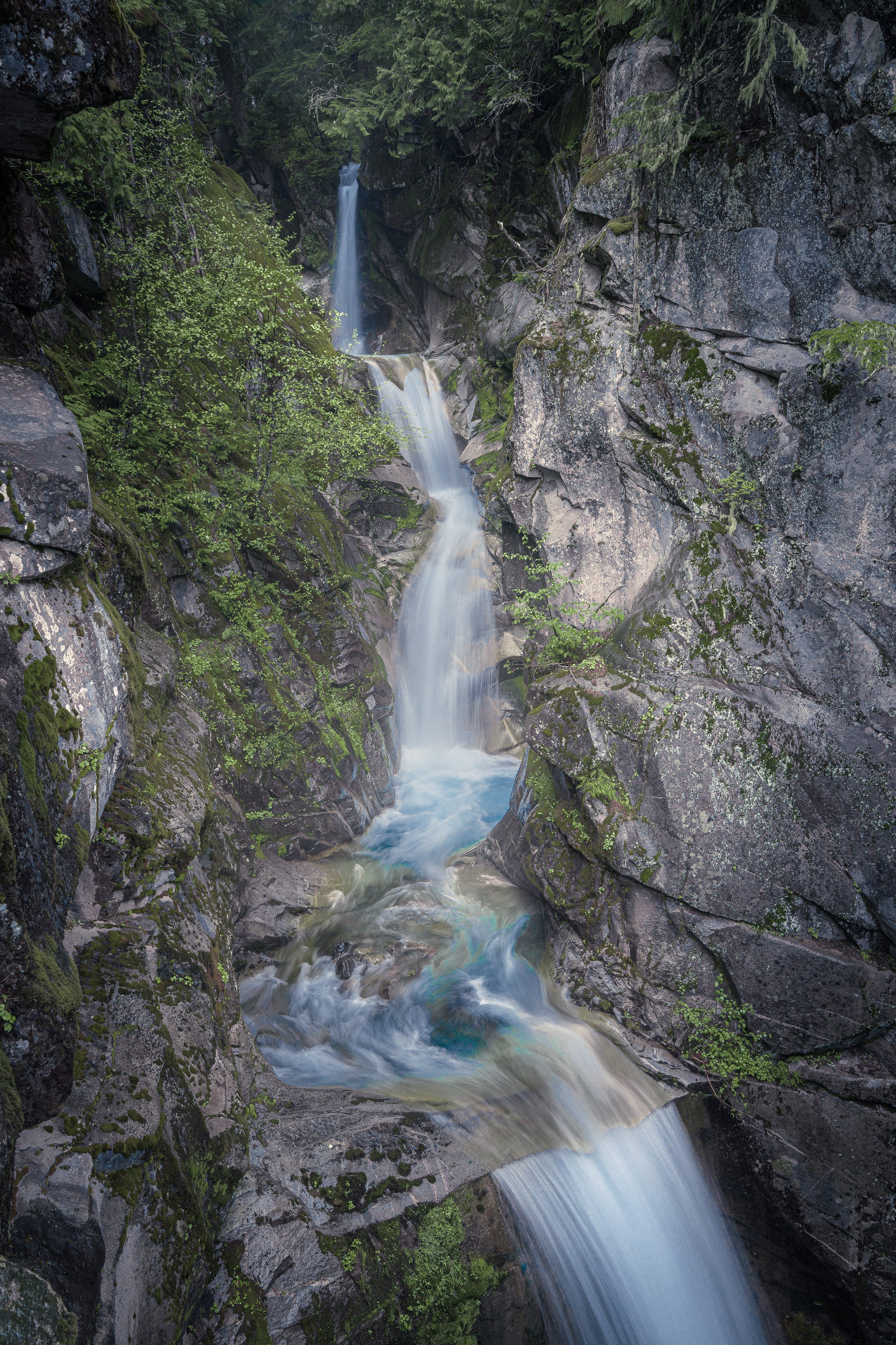 A multi-tiered waterfall flowing through a rocky canyon surrounded by dense green trees and moss-covered rocks.