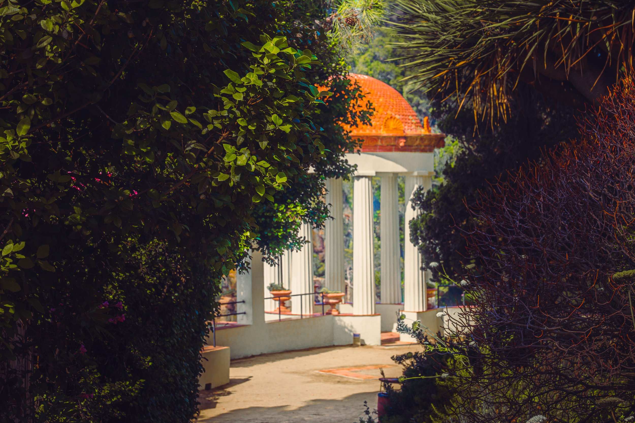 View through lush green foliage revealing a white building with classical columns and an orange tiled dome roof in the background.