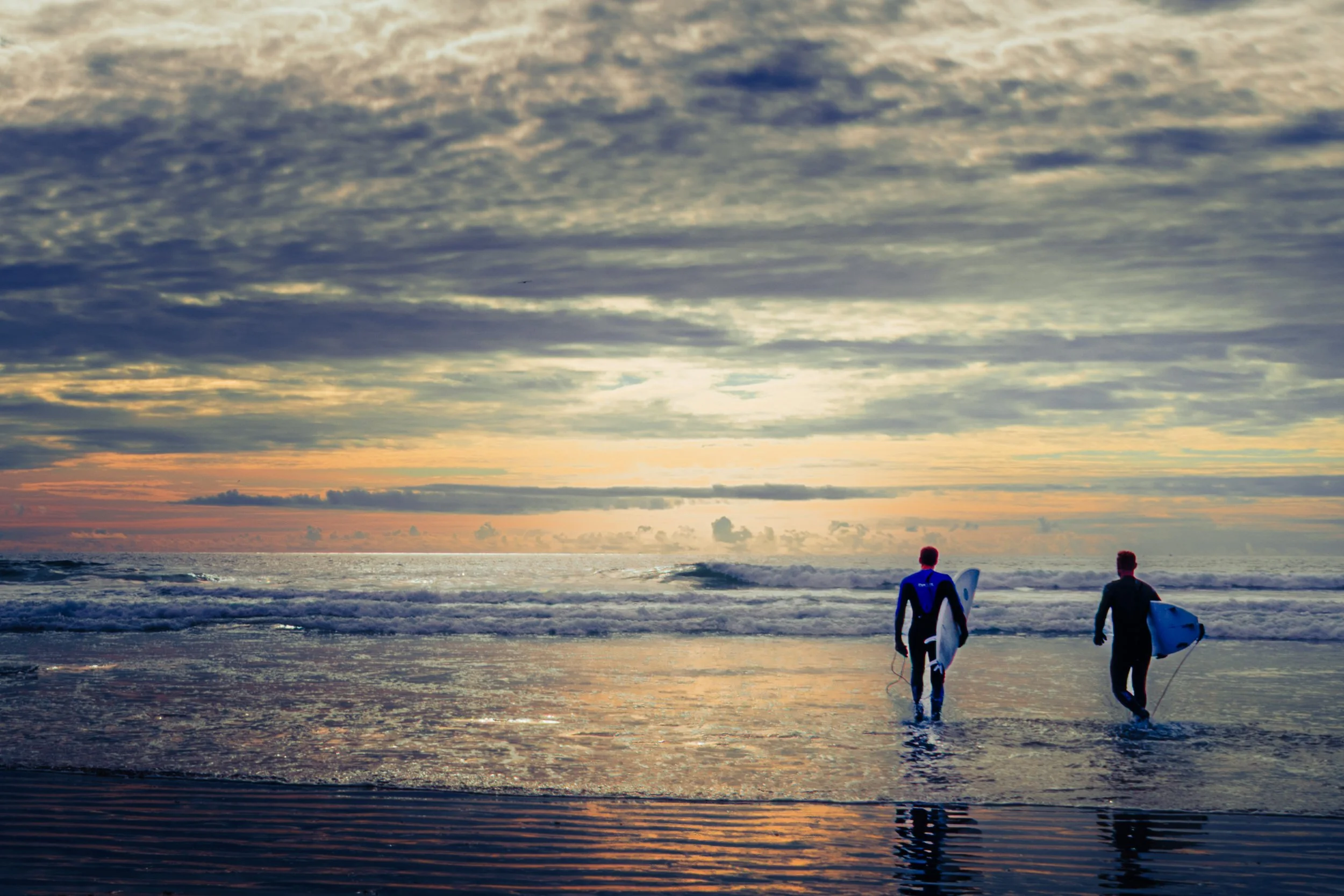 Two surfers walking into the ocean at sunset, carrying surfboards, with a cloudy sky overhead.