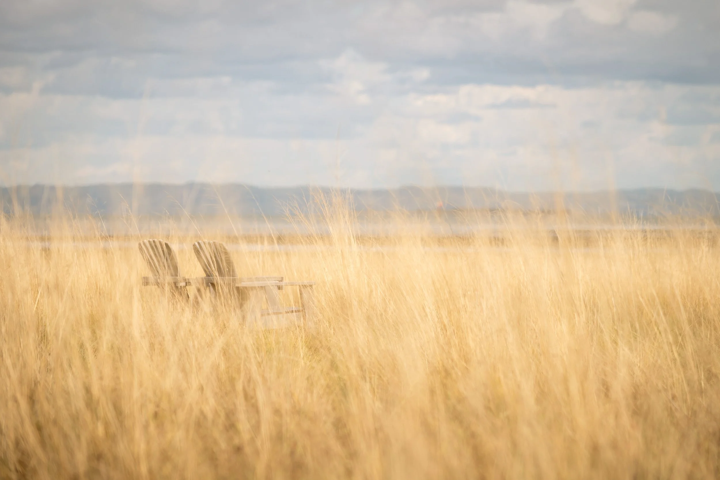 Two wooden chairs sitting on a grassy field with tall yellow grass, under a cloudy sky, with hills in the distance.