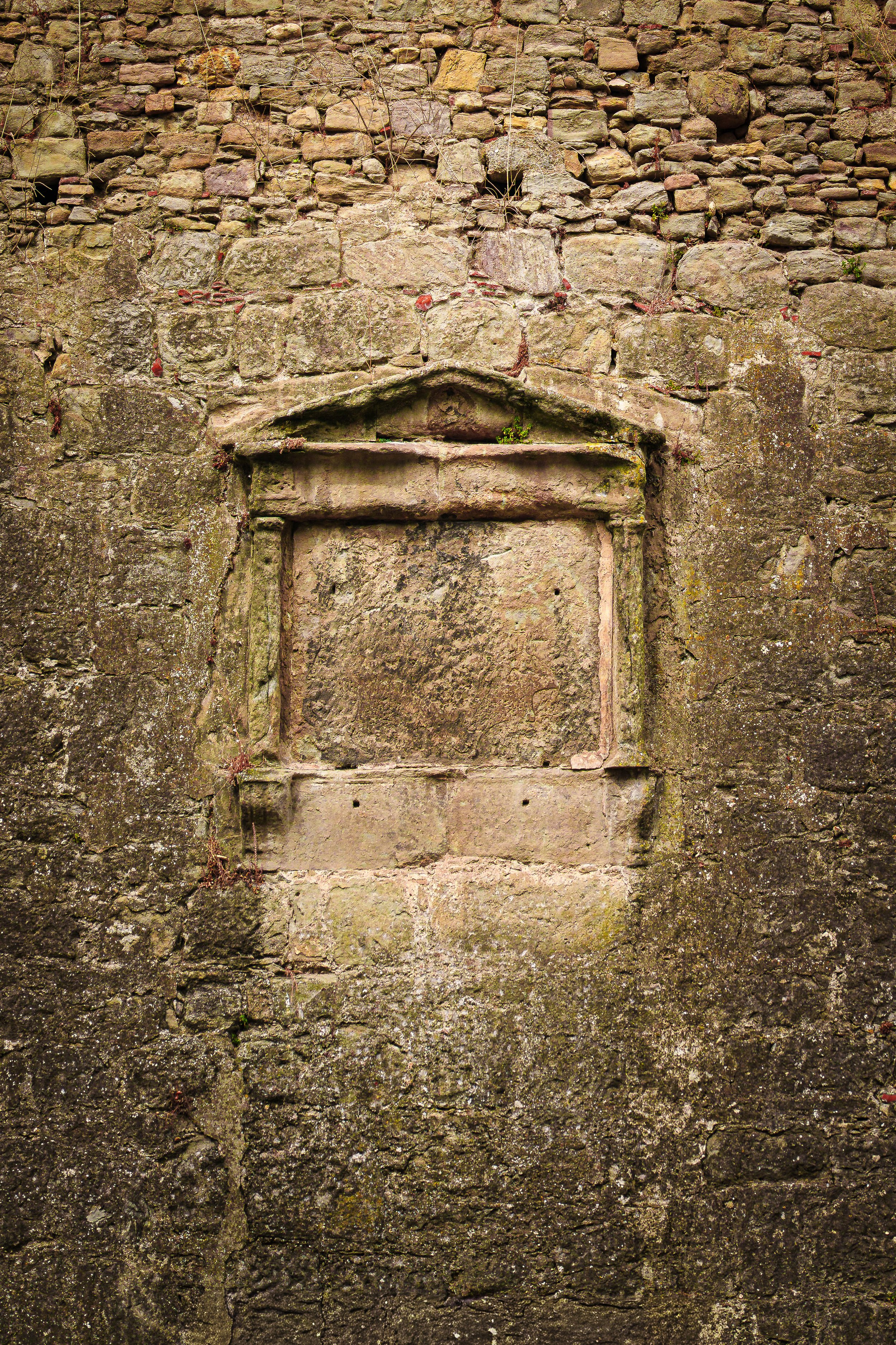 An old stone wall with an empty, recessed niche or frame at the bottom center, weathered with moss and small plants growing.