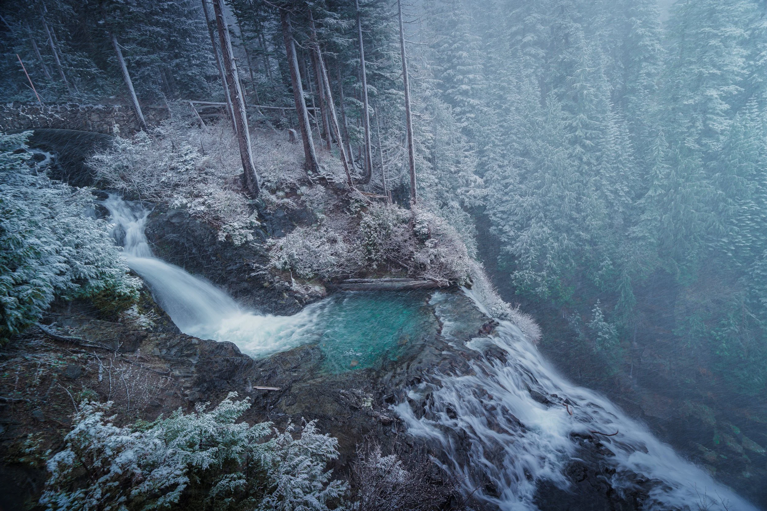 A long-exposure view looks across the upper cascades of Narada Falls toward a snowy evergreen forest fading into mist.