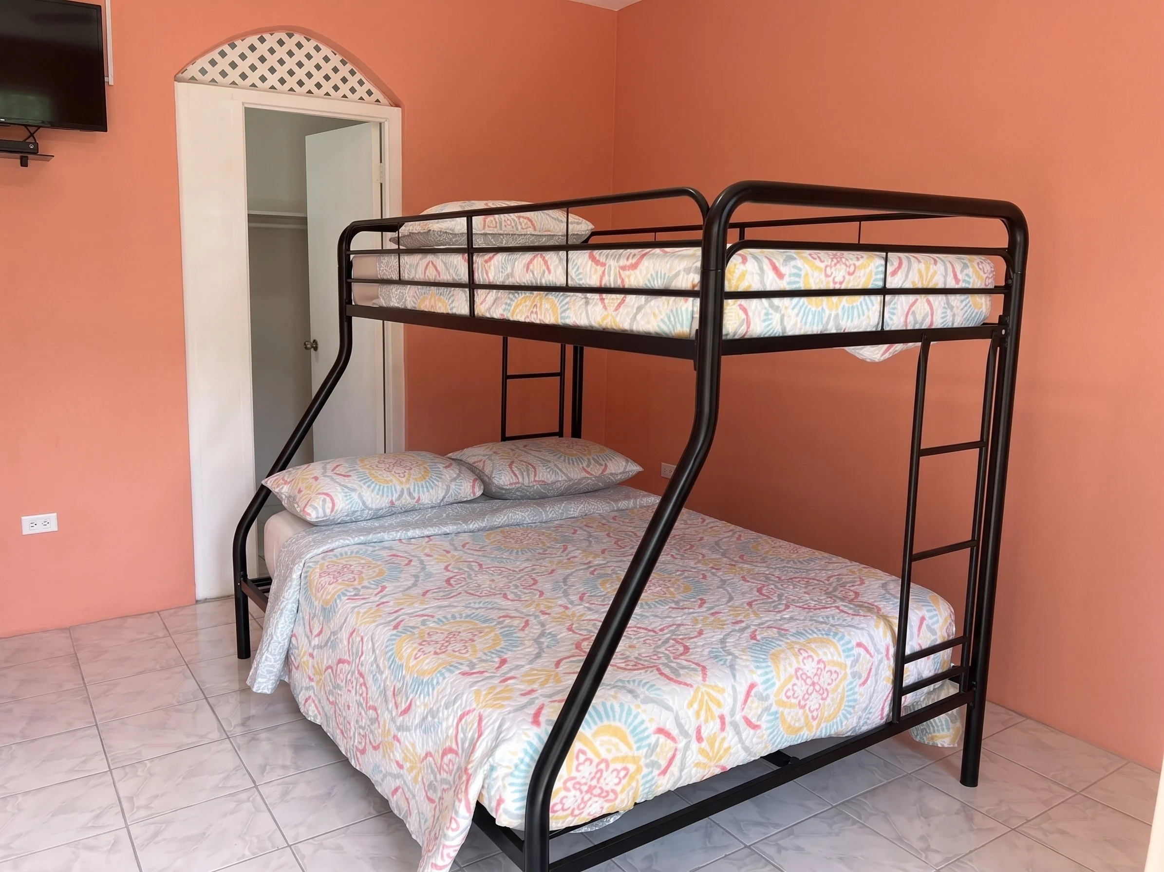 A room with peach-colored walls and tiled floor. A black metal bunk bed with floral-patterned bedding. The top bunk has pillows and bedding, and the bottom bunk is partially visible with similar bedding. There is a small closet with a white door in the corner and a wall-mounted TV on the left.
