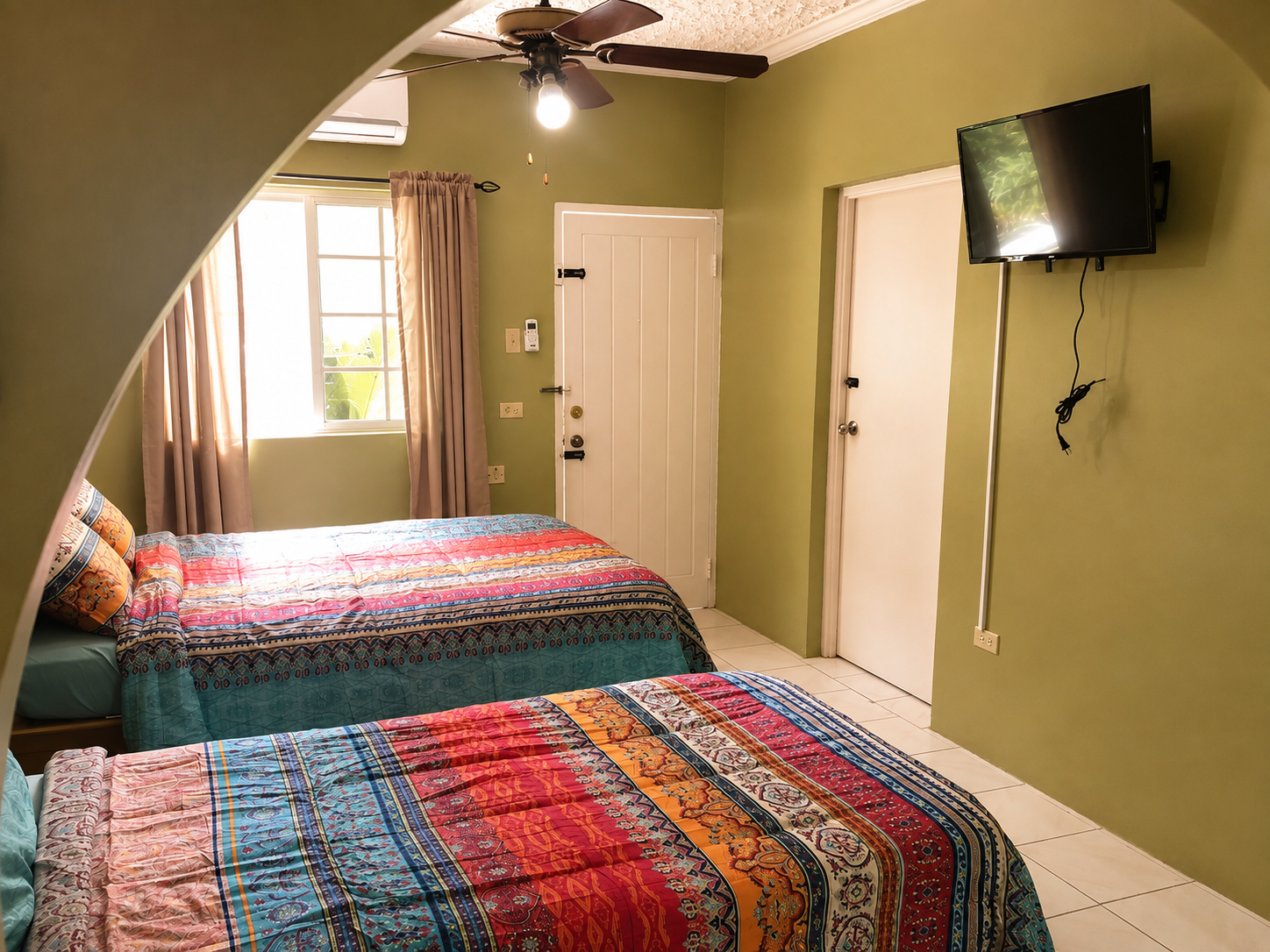 A bedroom with green walls featuring two beds with colorful patterned bedspreads. A window with curtains lets in natural light, and a ceiling fan with a light is mounted on the ceiling. There is a small wall-mounted television on the right wall and a closed door next to it.