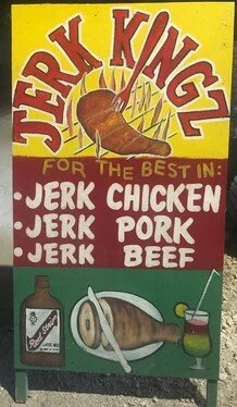 A colorful sign advertising jerk chicken, jerk pork, and jerk beef, with a drumstick illustration at the top, a bottle of hot sauce, a ham, and a drink with a straw at the bottom.