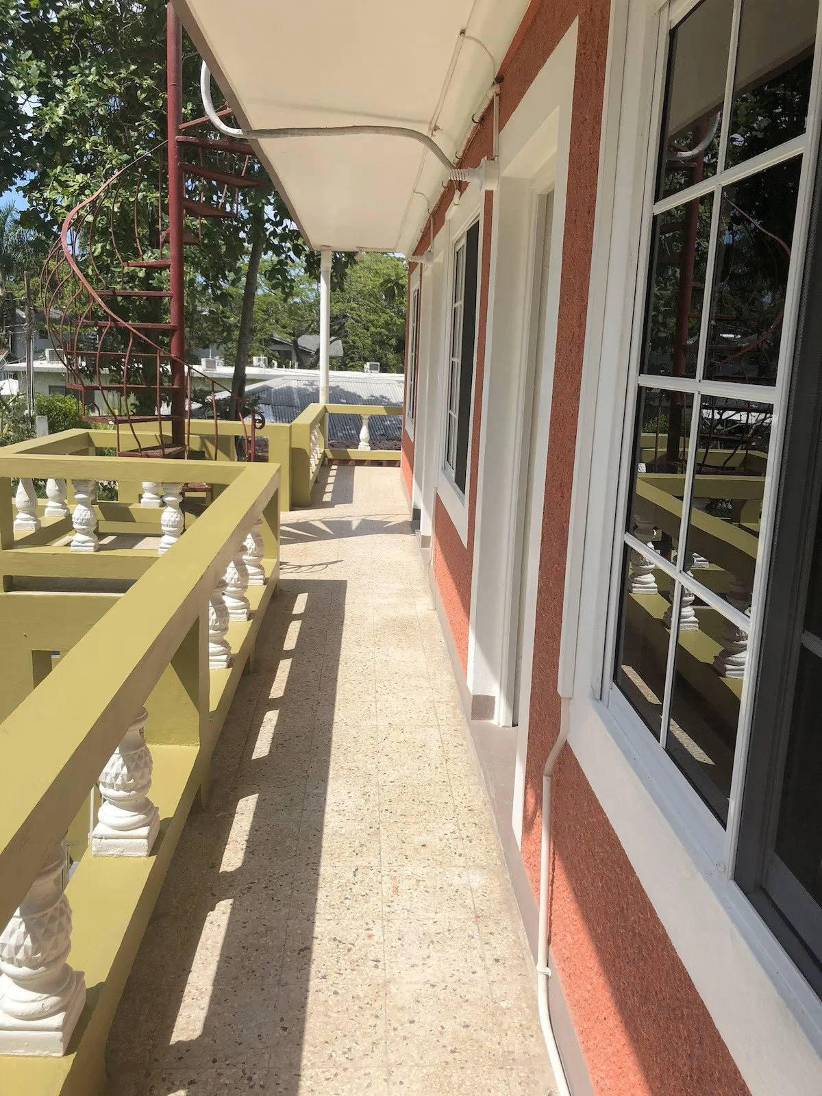 A balcony with yellow railing, red painted walls, and large windows reflecting greenery, on a sunny day with clear sky.