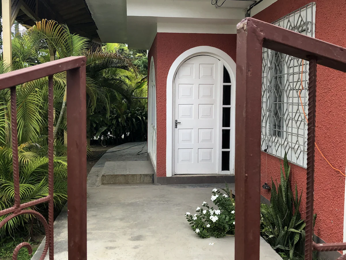 Front view of a house with a white arched door, a small step, a window with security bars, and a garden with green plants and white flowers, seen through a rusty metal fence gate.