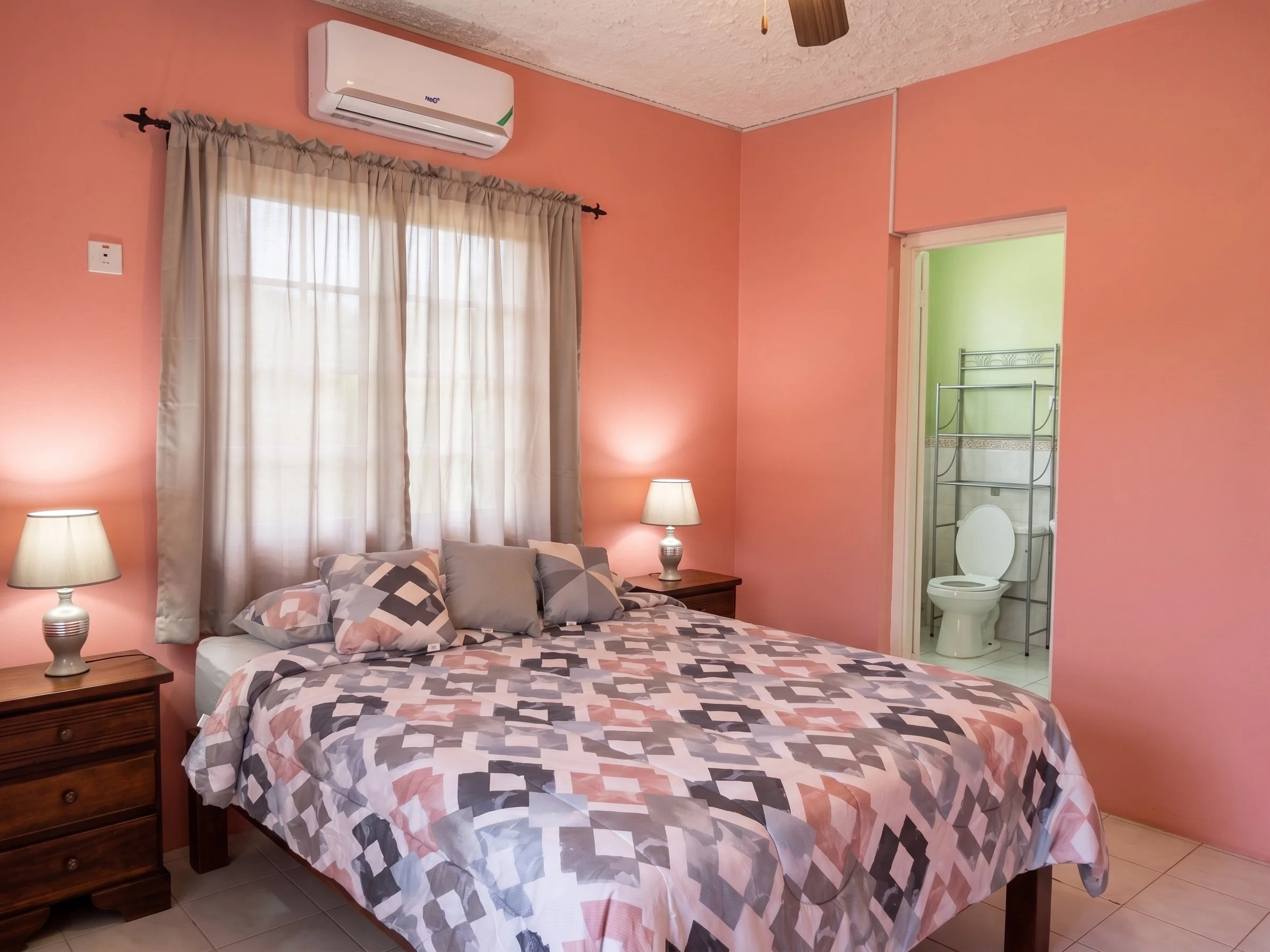 A bedroom with pink walls, a window with beige curtains, a bed with geometric patterned bedding, a wooden nightstand with a lamp, and a bathroom with a toilet visible through an open door.