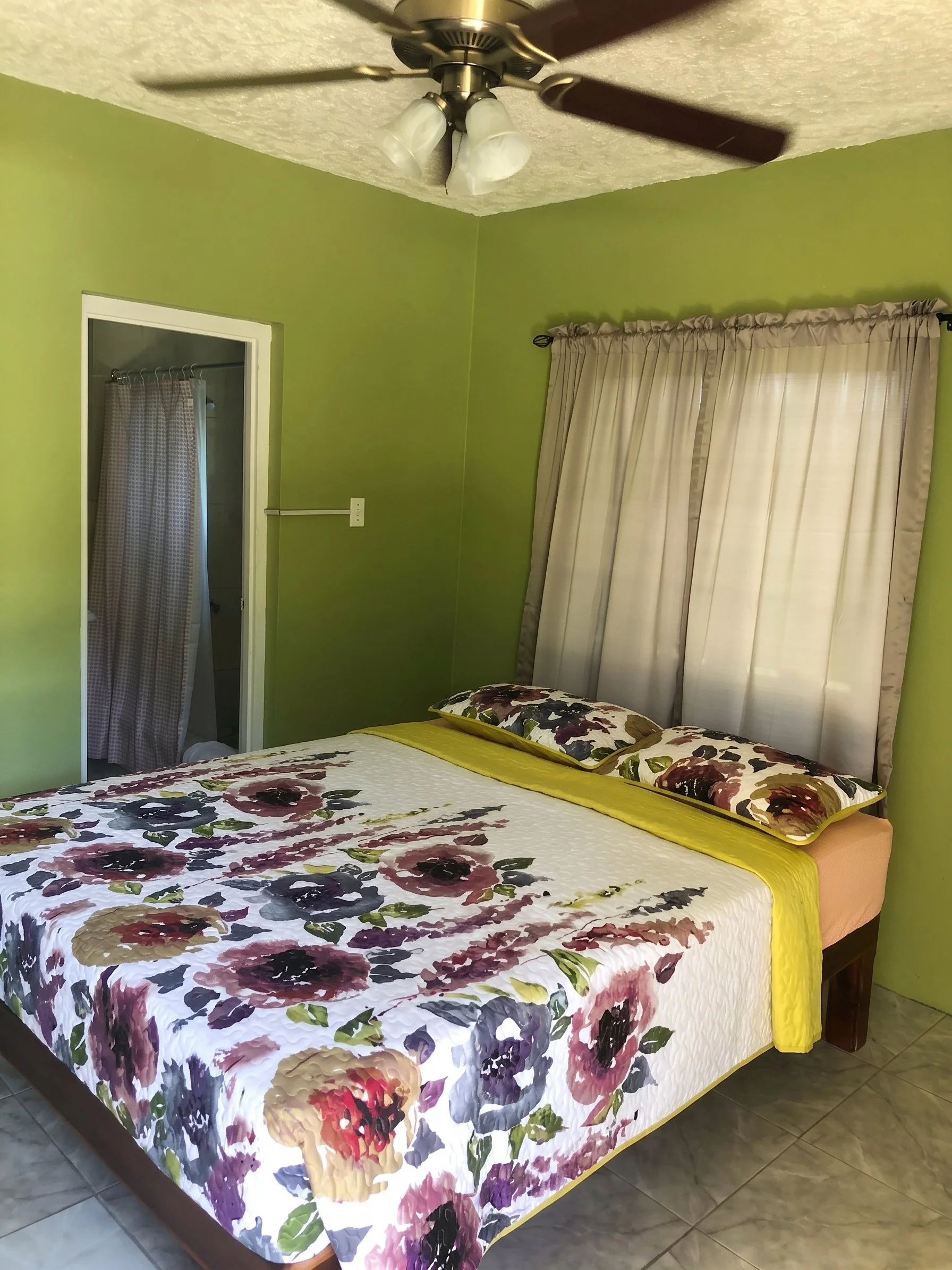 A bedroom with green walls and a floral-patterned bedspread, two pillows, a window with beige curtains, and an overhead ceiling fan with three lights.