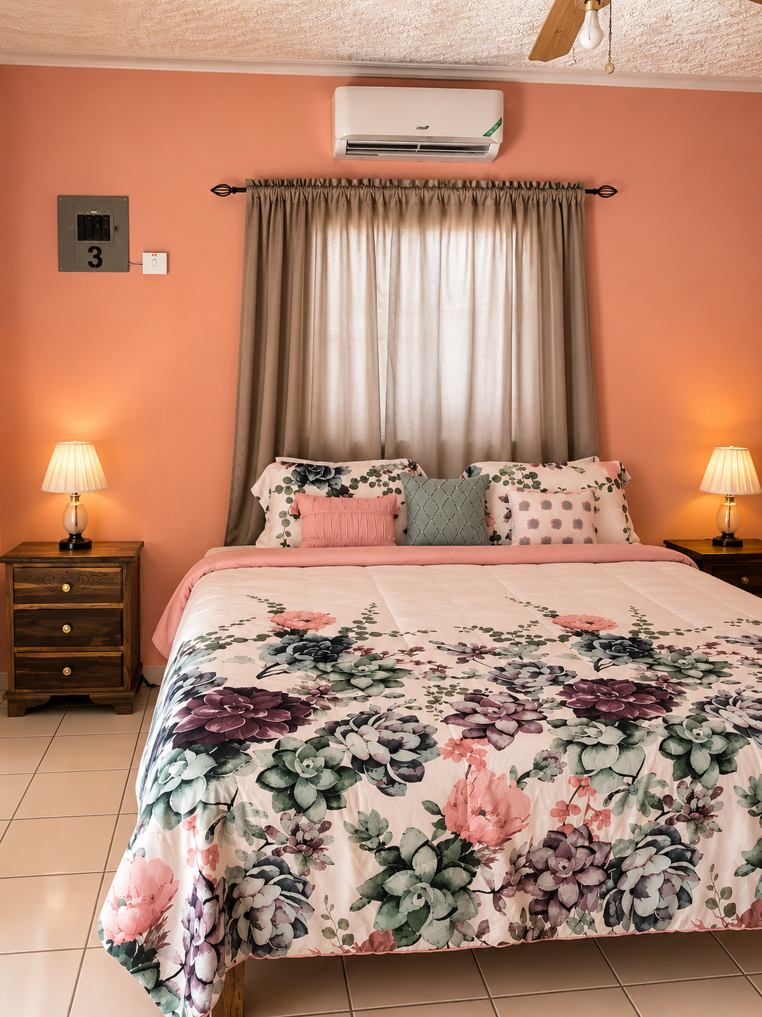 A bedroom with pink walls, a bed with floral bedding and pillows, beige curtains, two wooden nightstands with lamps, bamboo ceiling fan, and a wall air conditioning unit.