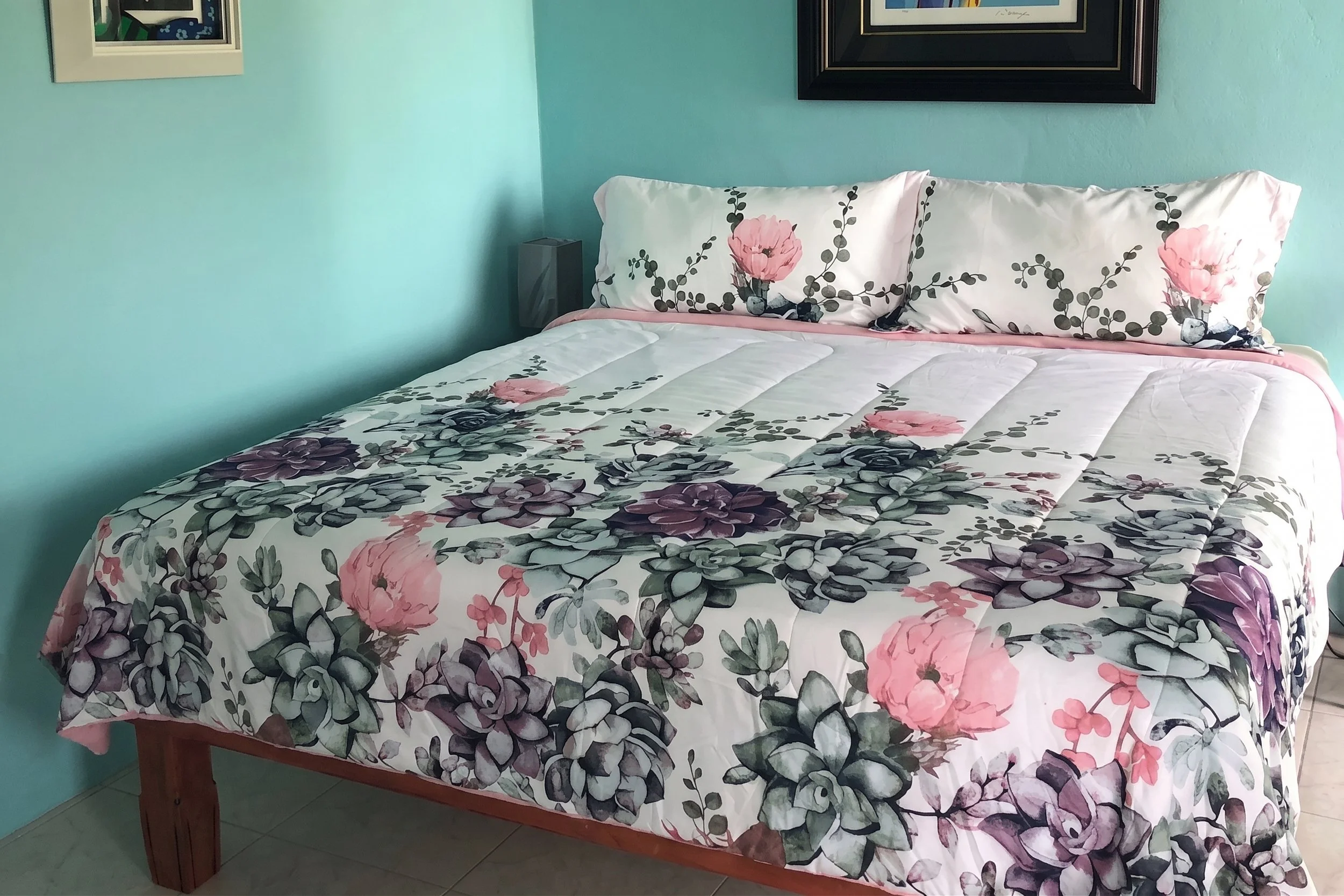 A bed with a floral bedspread and pillowcases featuring pink and purple flowers and green leaves, set against a light blue wall with framed pictures hanging above.