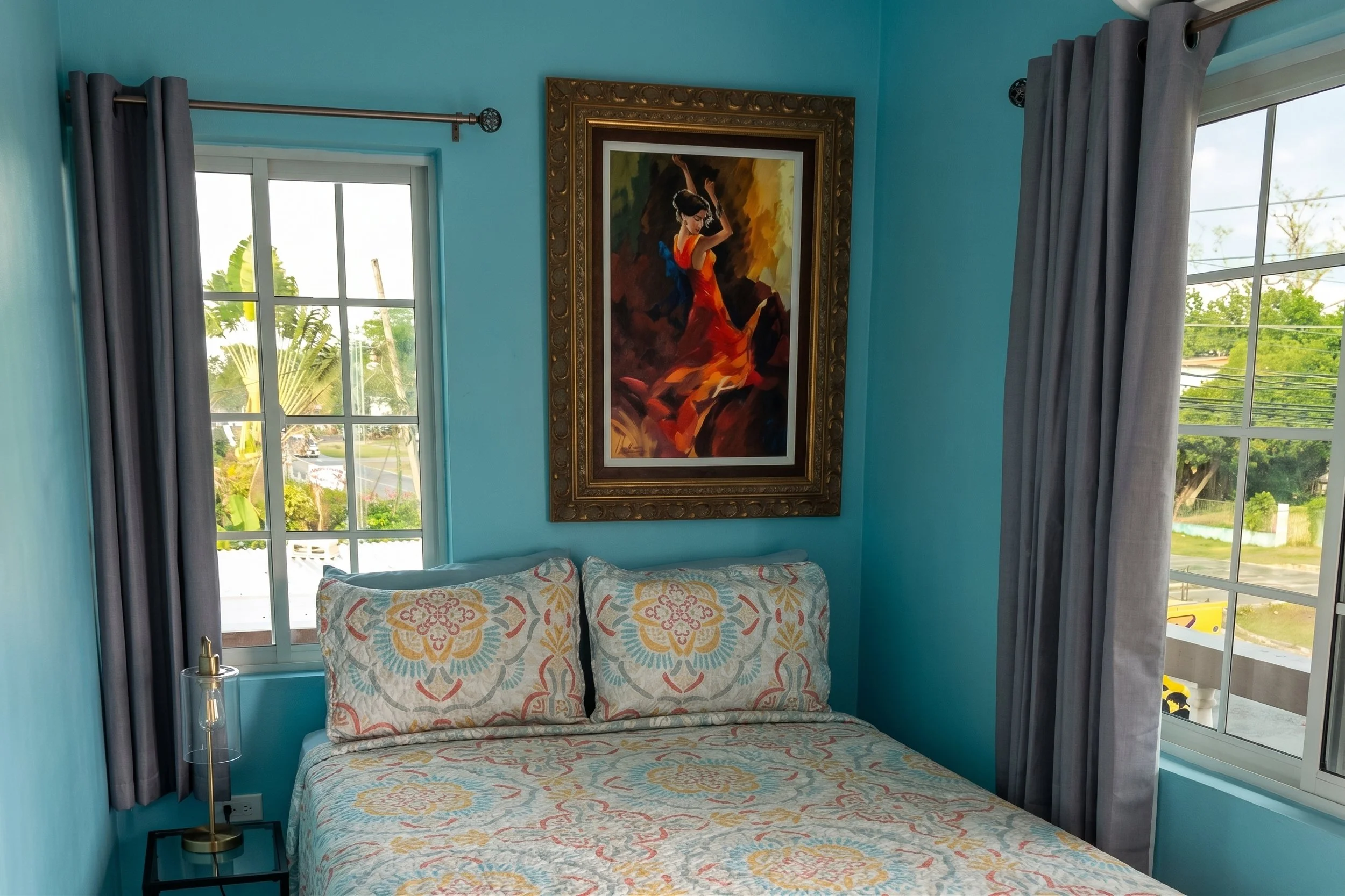 A bedroom with teal walls, two large windows with silver curtains, a bed with a patterned quilt, a framed painting of a woman in a red dress, and a small bedside table with a clear lamp.