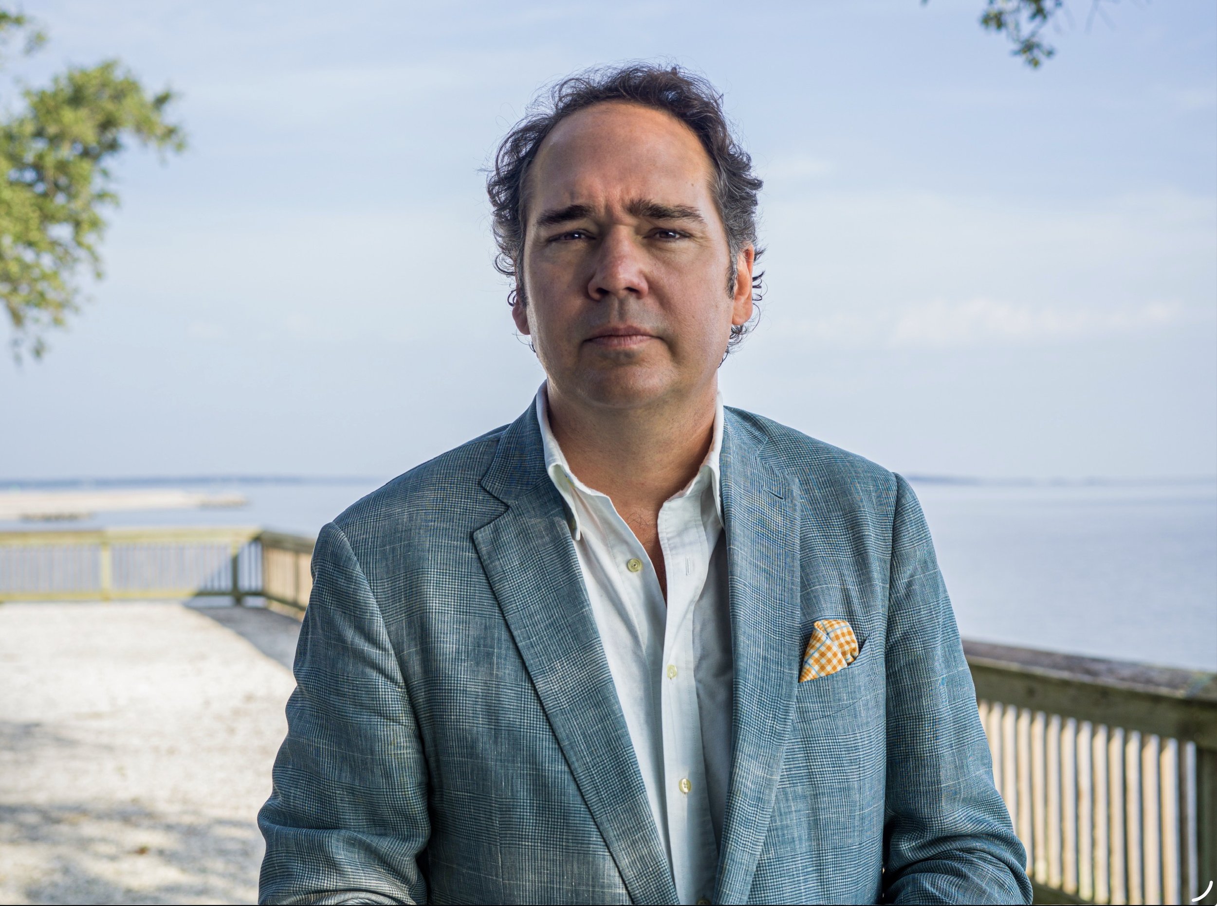 A man in a gray checkered blazer and white shirt standing outdoors near water, with trees and a wooden fence in the background.