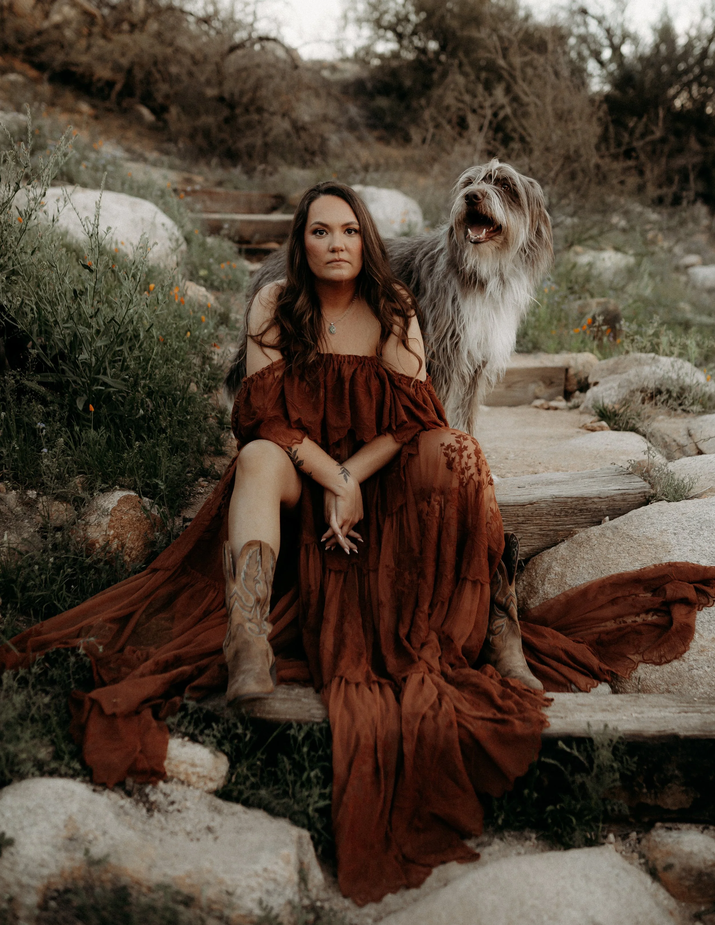 A woman with long brown hair wearing an off-shoulder rust-colored dress sitting on a rock in a natural outdoor setting with greenery and rocks, posing with a large fluffy dog standing beside her.