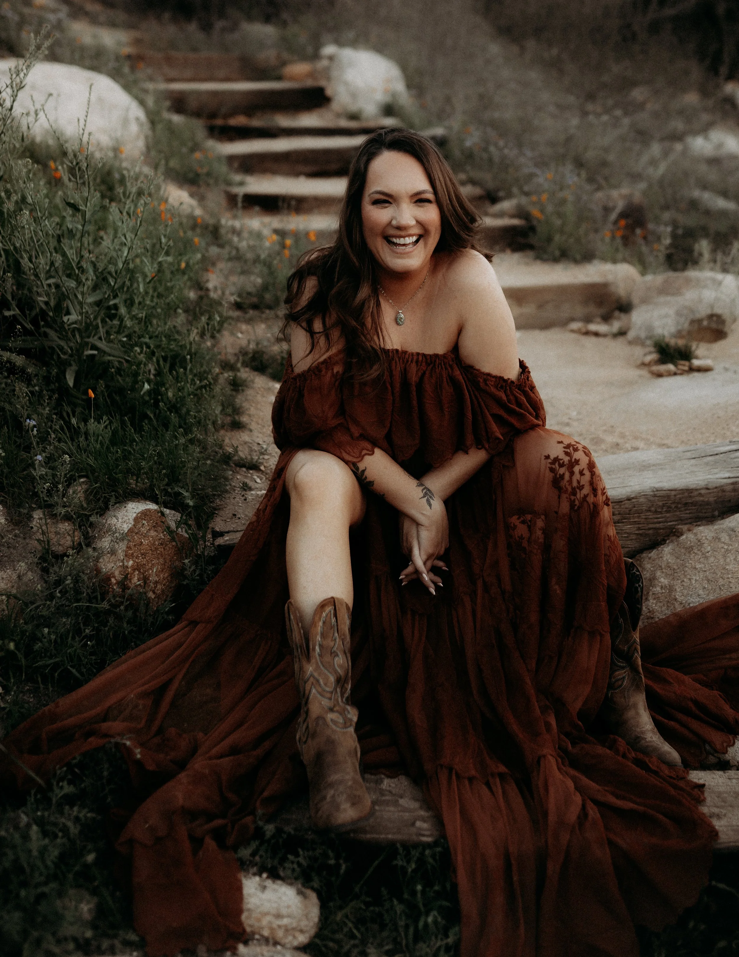 A woman with long dark hair smiling and sitting outdoors on a rocky trail surrounded by desert plants, wearing a dark red off-shoulder dress and cowboy boots.