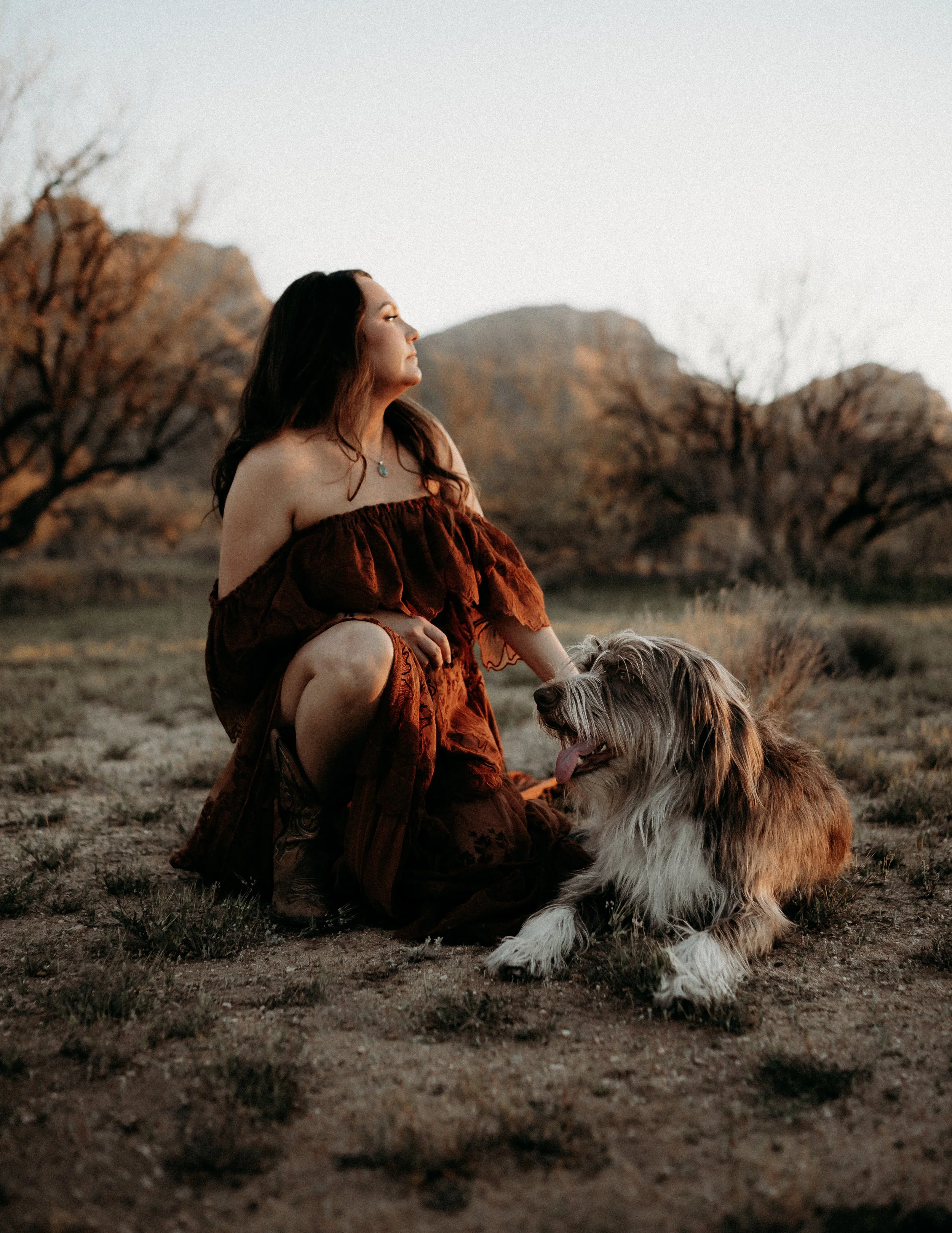 A woman with long hair wearing a reddish-brown off-shoulder dress sitting on the ground with a dog in a desert landscape with mountains in the background at sunset.