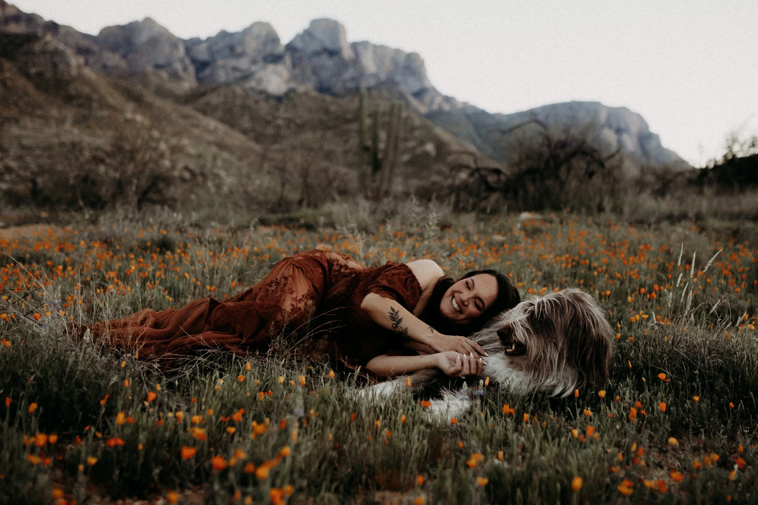 A woman lying on the ground in a field of orange flowers, smiling and petting a fluffy dog, with mountains in the background.