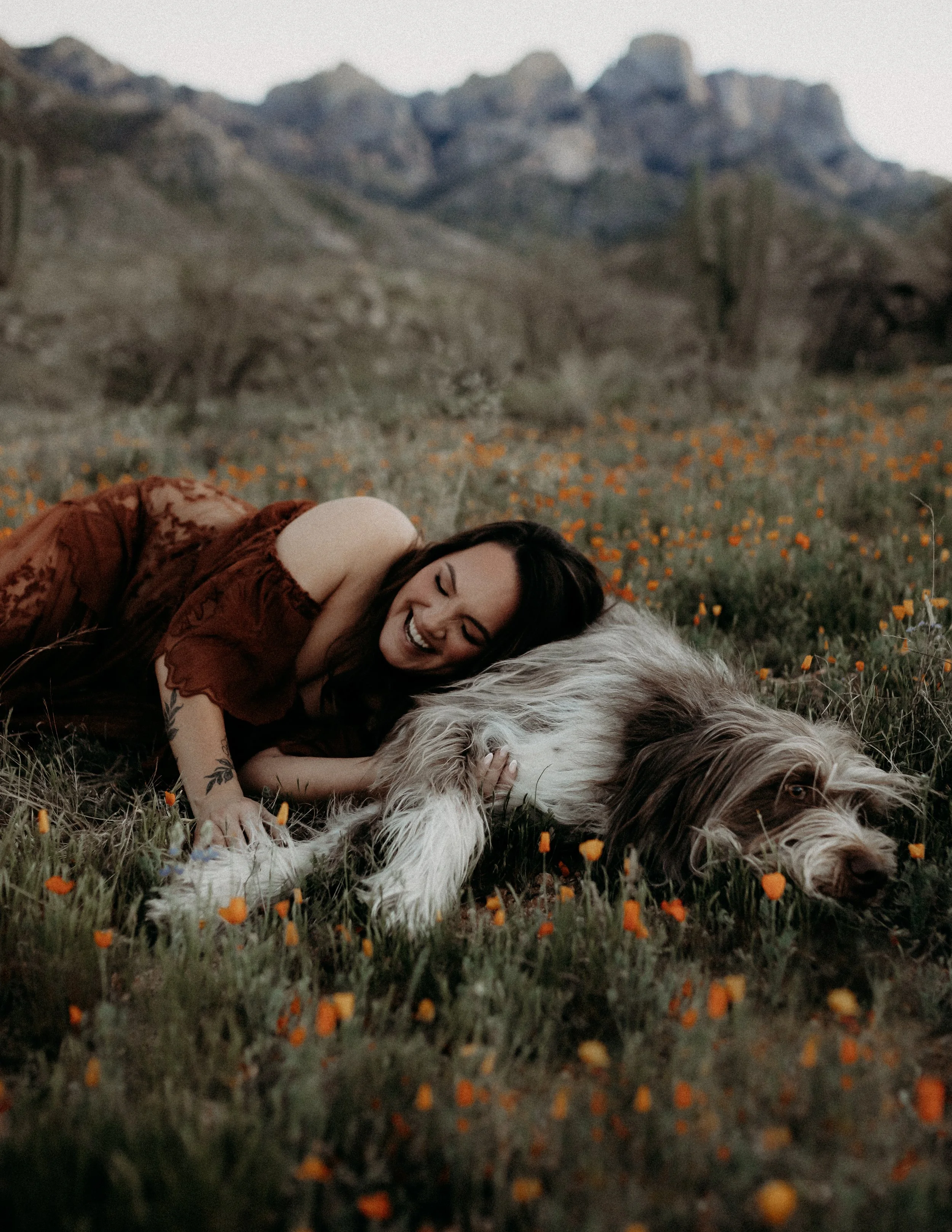 A woman lying on grass among orange flowers in a field with mountains in the background, smiling and cuddling a large dog.