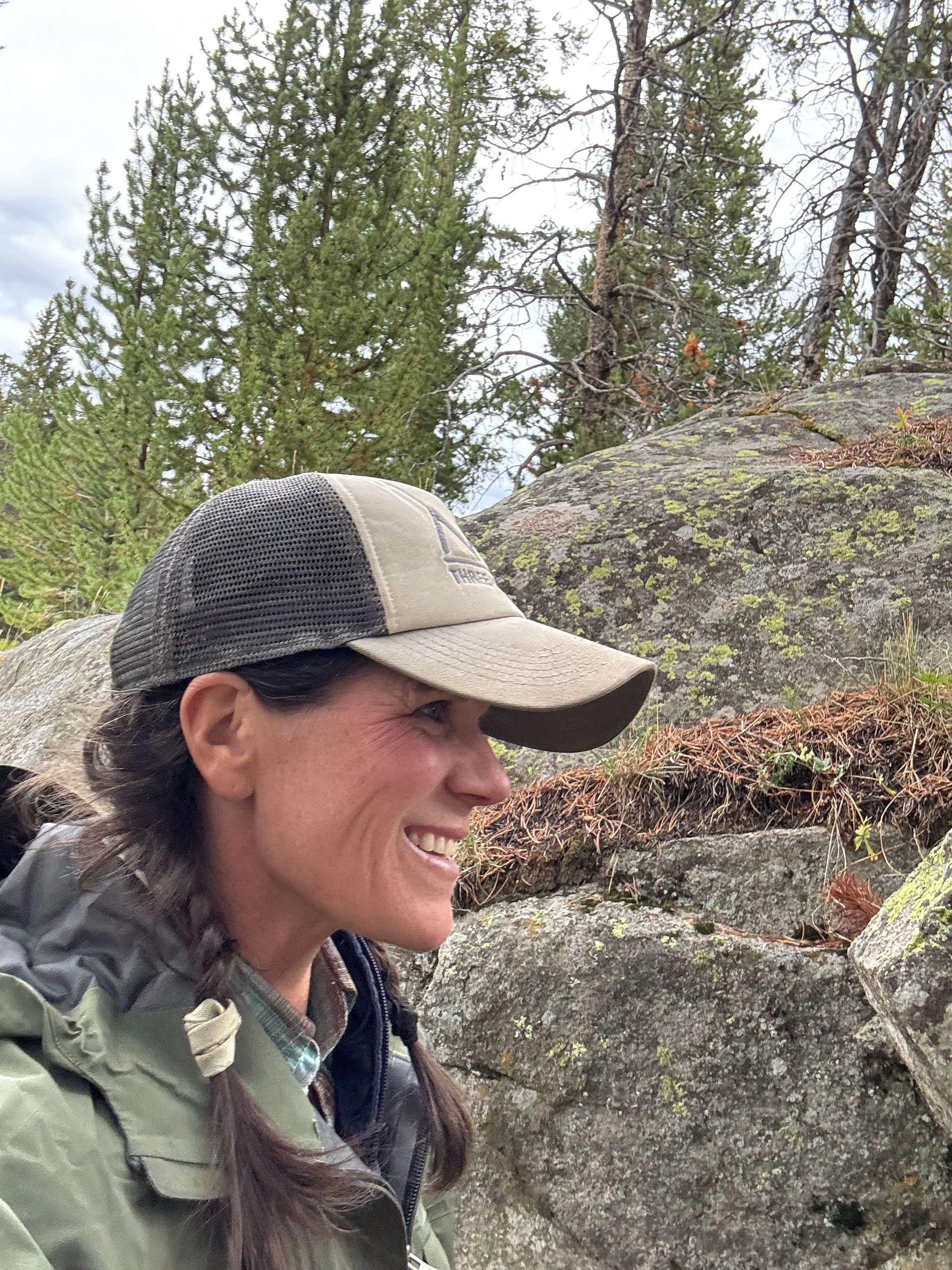 A woman with long brown hair wearing a beige cap, green jacket, and backpack smiling while outdoors among rocks and trees in a forested area.
