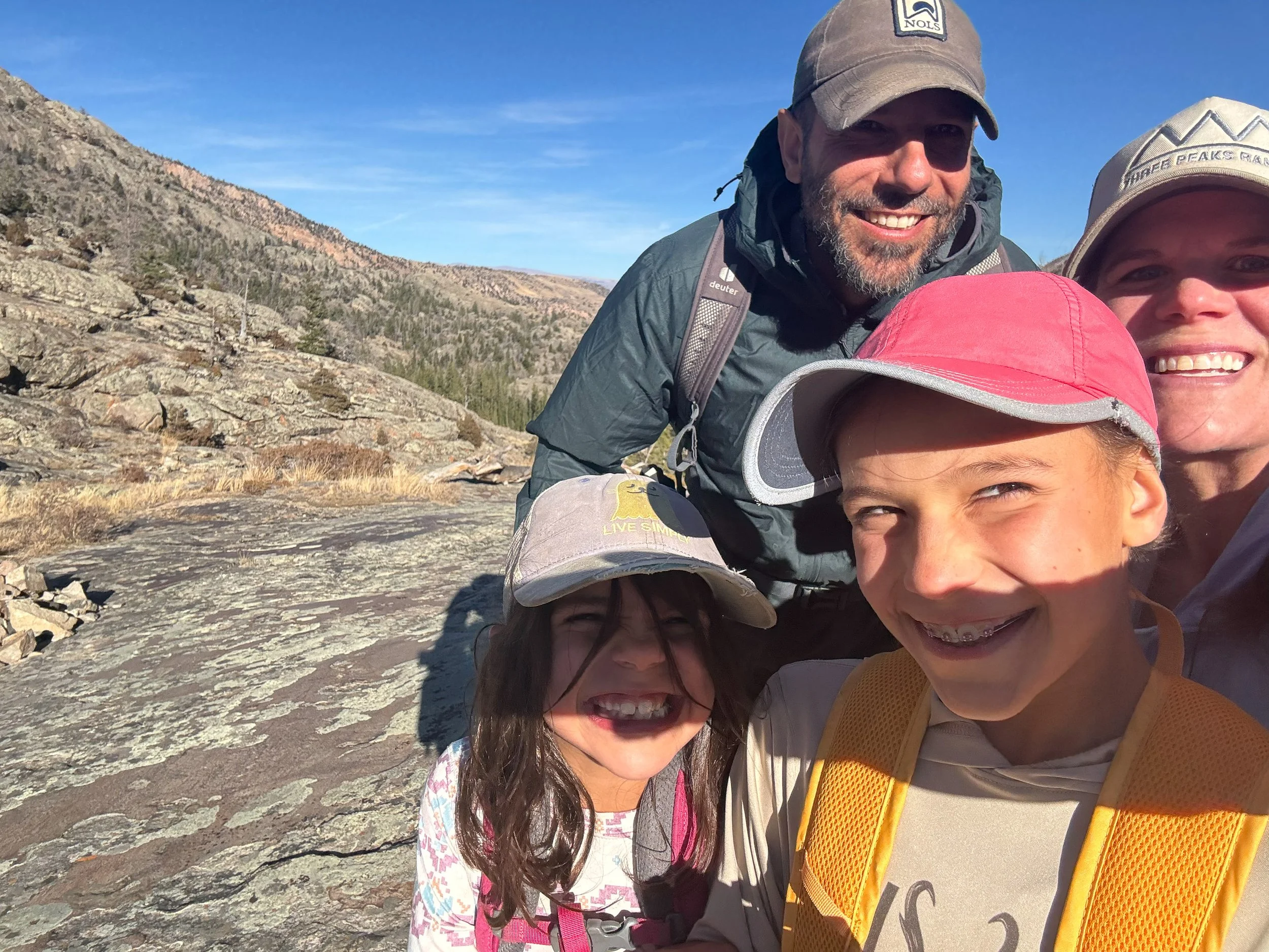 A group of smiling hikers taking a selfie on a rocky mountain trail under a clear blue sky with a mountain range in the background.