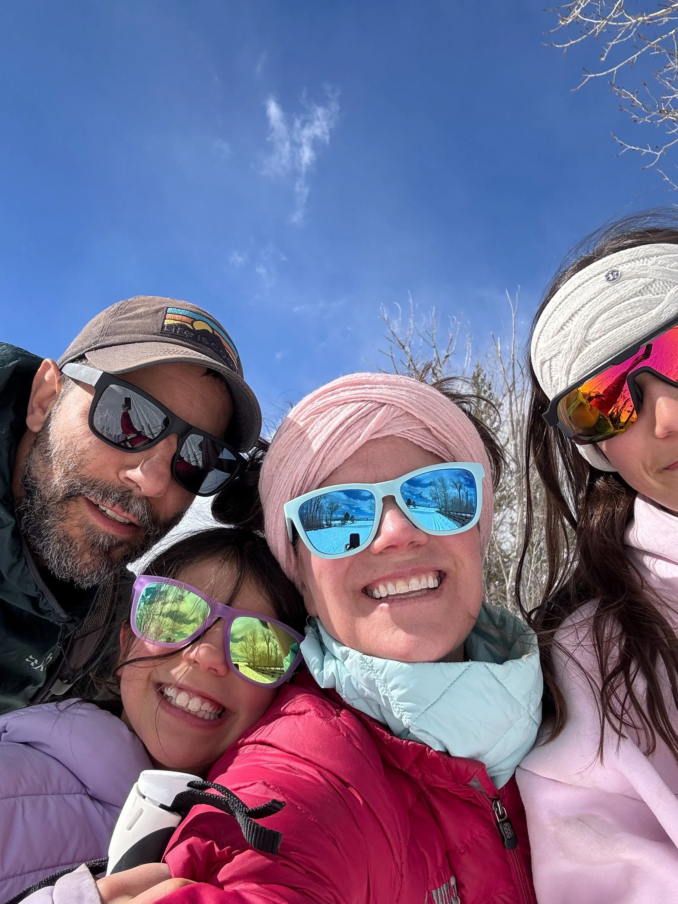 Group of four people taking a selfie outdoors under blue sky, wearing sunglasses, hats, and jackets, with clear weather and bare trees in the background.