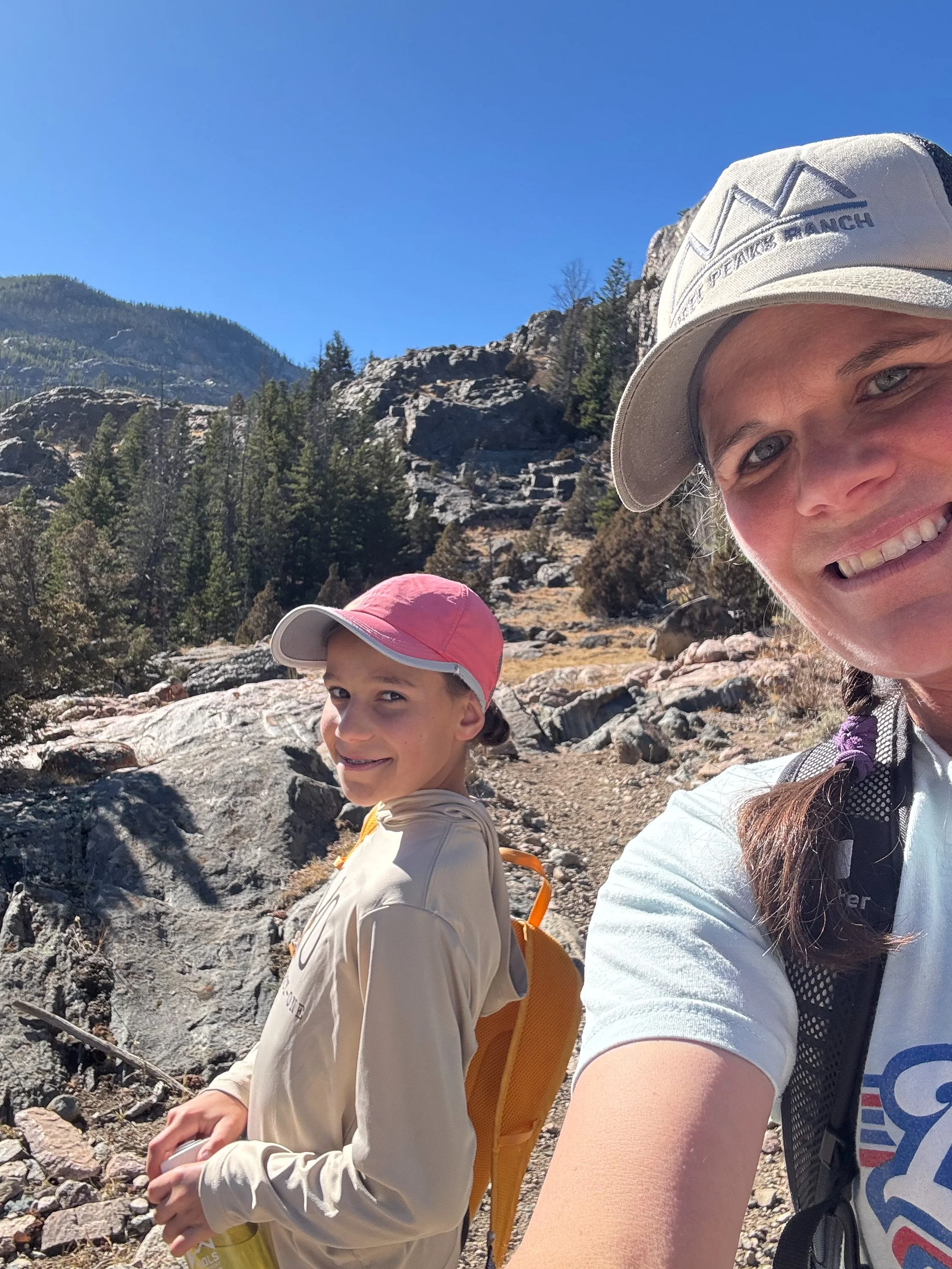 A woman and a girl are hiking on a rocky trail with mountains, trees, and a clear blue sky in the background. The woman is smiling, wearing a light-colored hat and a white shirt. The girl is looking back, wearing a pink hat and a beige jacket. Both h