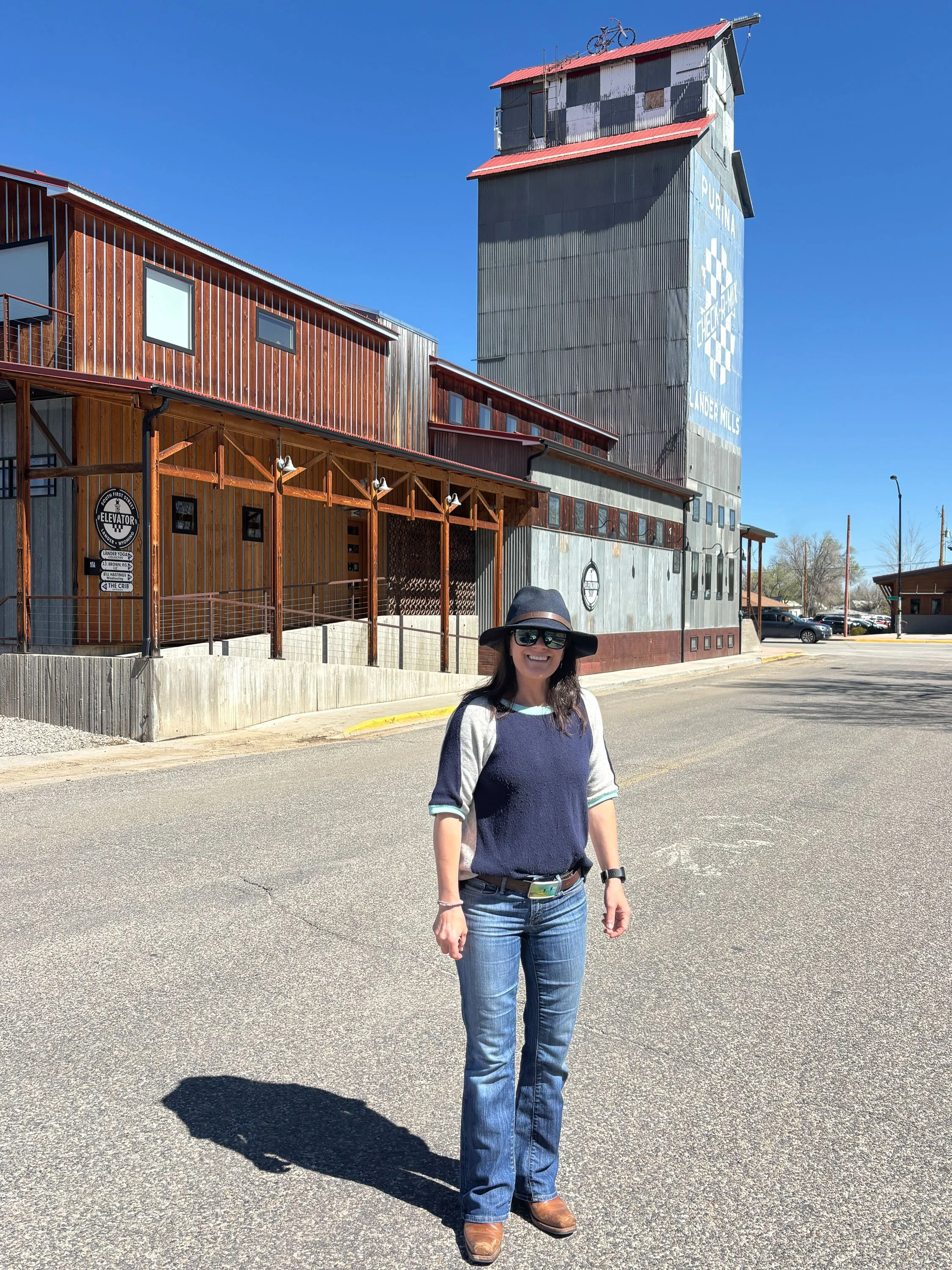 A woman smiling, wearing sunglasses, a wide-brimmed hat, a navy and gray long-sleeve shirt, jeans, and boots, standing on a paved road with a large industrial building in the background. The building has a tall silo with a bicycle on top and a sign t