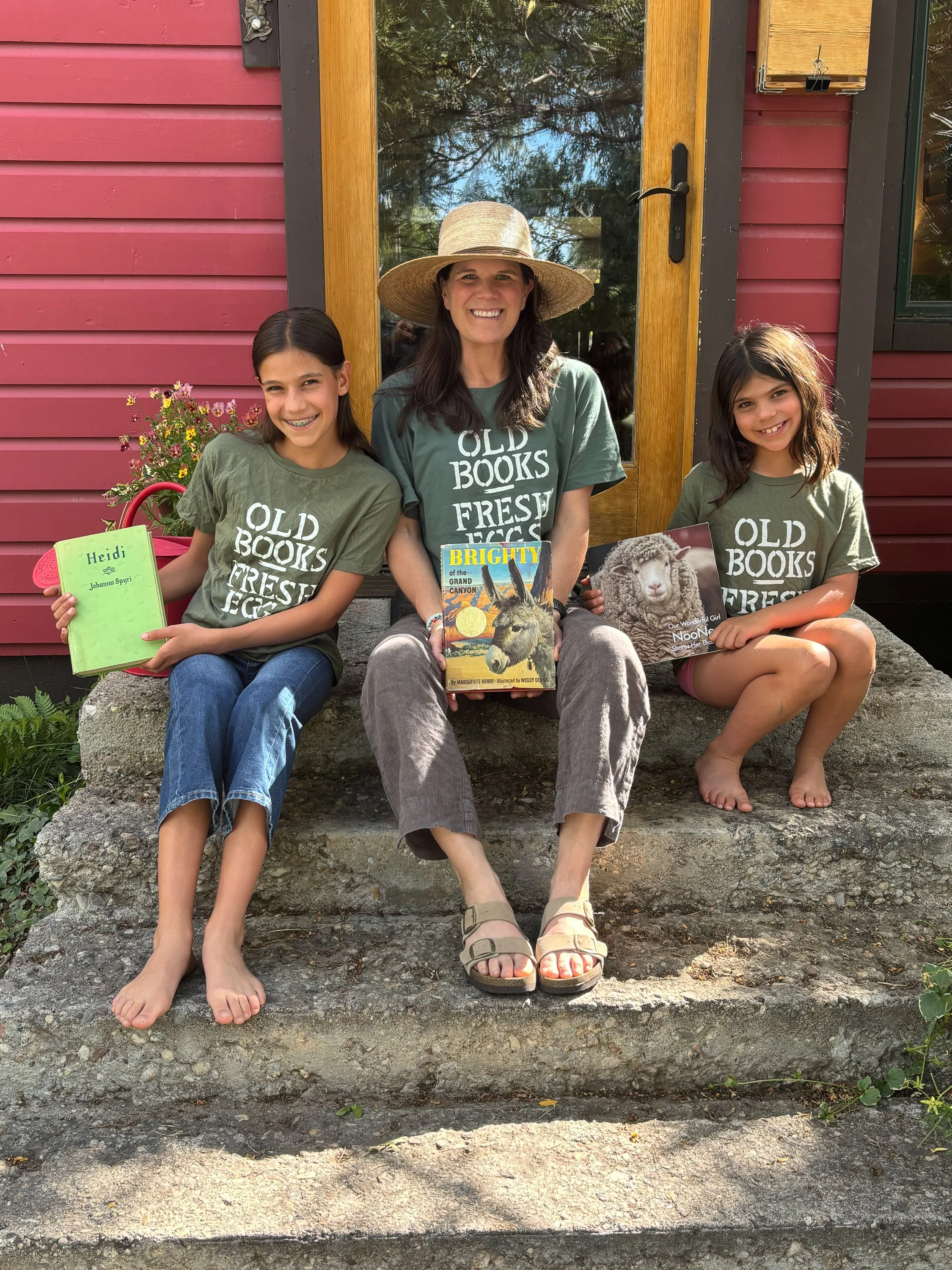 A woman and two girls sitting on a stone step outside a red house with a wooden door and window. The woman is in the middle wearing a large straw hat, a dark green T-shirt, and gray pants, holding a colorful book. The girl on the left has dark hair i