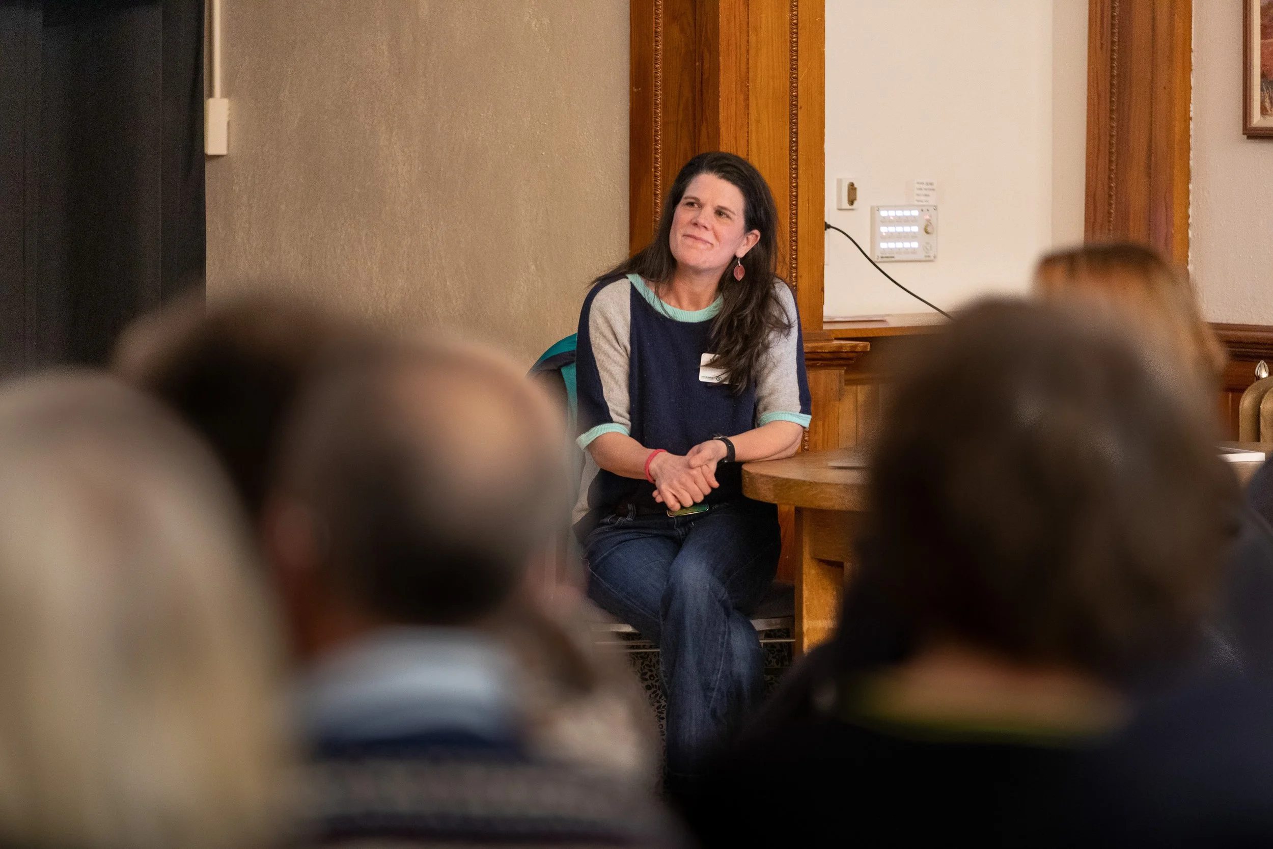 A woman with dark hair and earrings sitting at a wooden table, speaking or listening during an indoor event with multiple people, some of whom are visible in the foreground, and a beige wall with wooden accents and a control panel in the background.