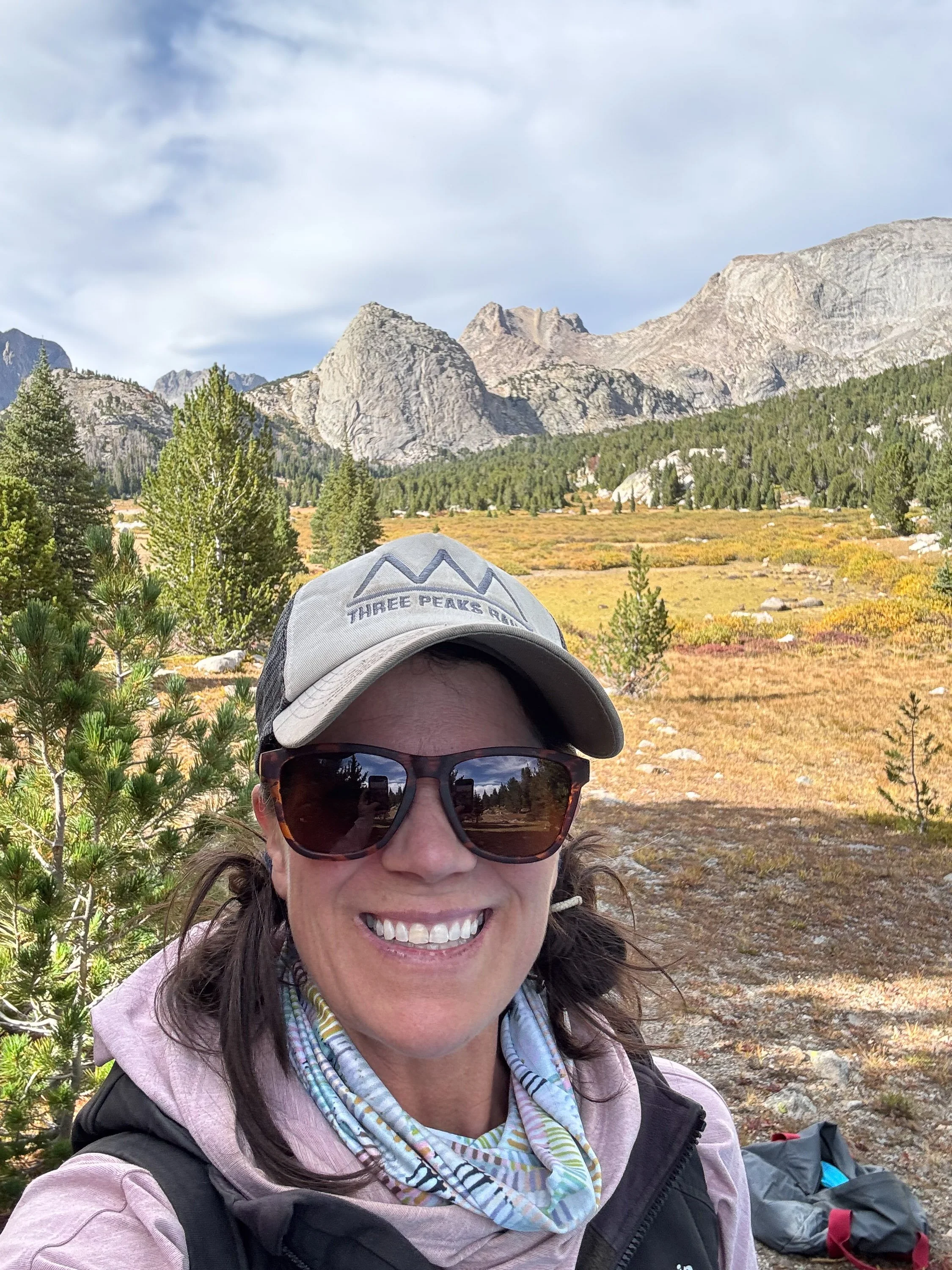 A woman smiling outdoors in a mountainous landscape with trees and rocky peaks in the background, wearing sunglasses, a cap, and a jacket.