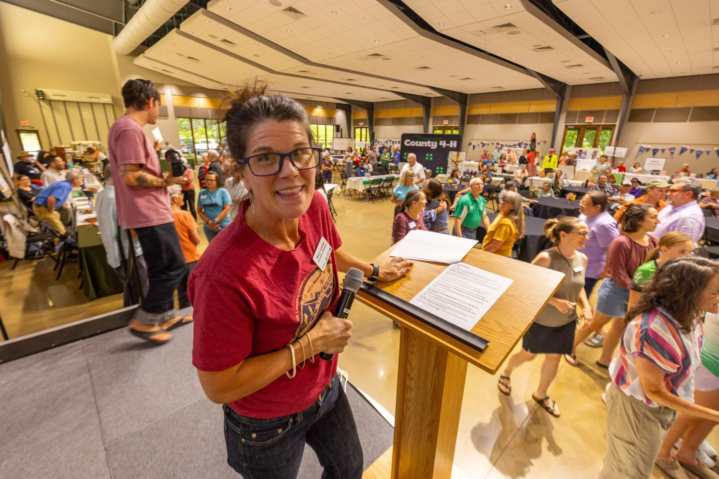 A woman speaking at a community event in a large indoor hall filled with people. She is wearing glasses, a red shirt, and holding a microphone, standing behind a wooden podium with documents. The hall is decorated with banners and booths, and a crowd