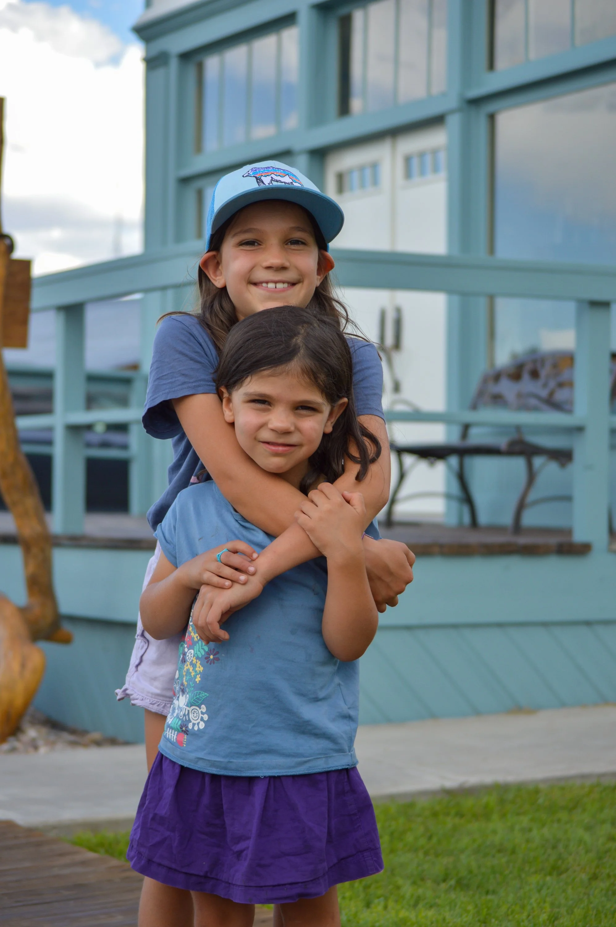 Two young girls hugging in front of a light blue house with large windows and a porch, smiling outdoors.